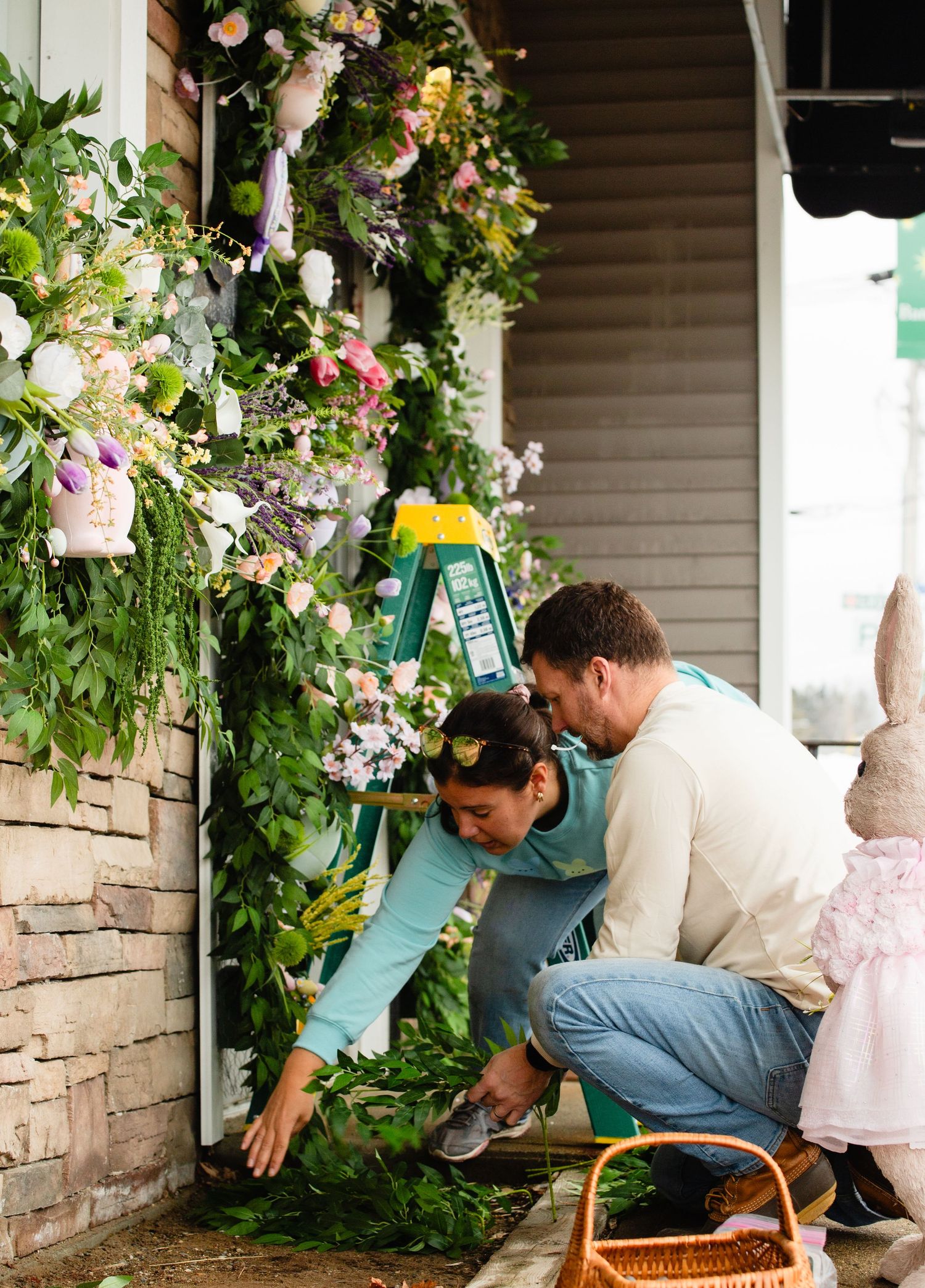 Father and child kneeling together arranging green garland on a flower-decorated porch.