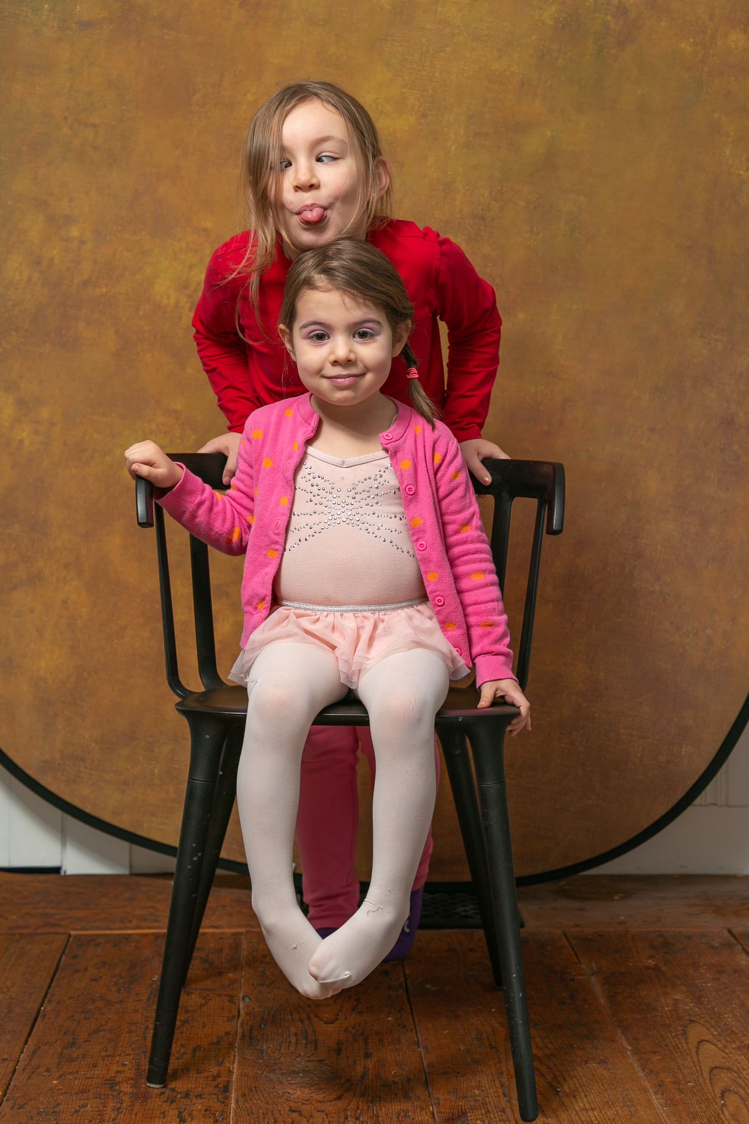 Two young girls posing playfully with a black chair against a brown studio background.