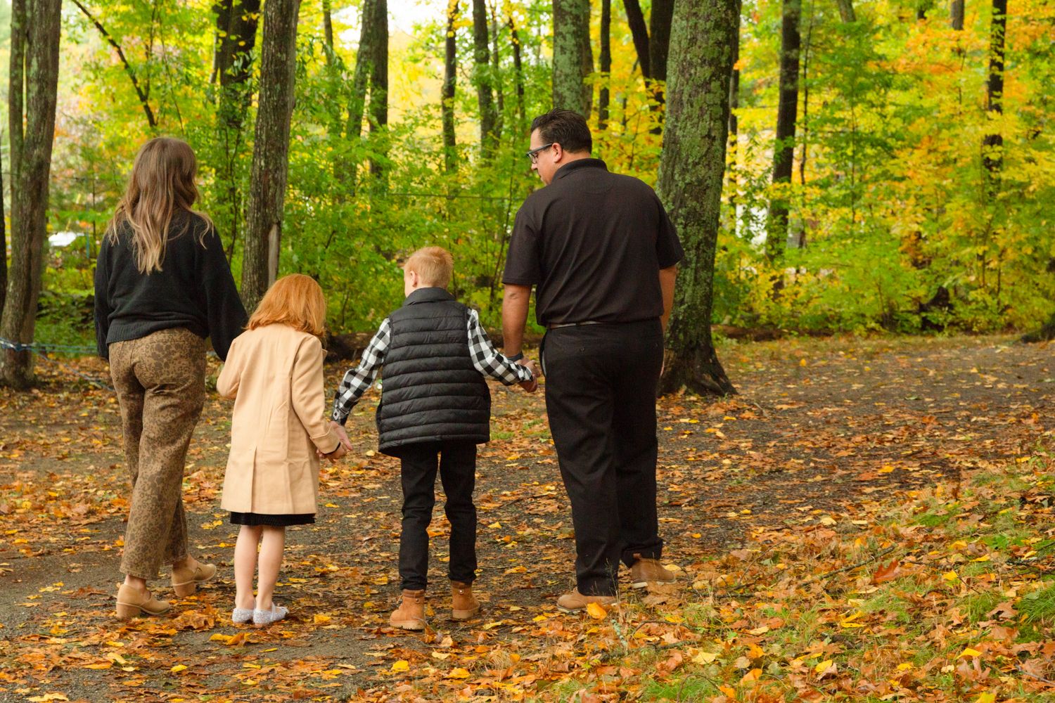 A family of four holds hands while walking along a leaf-covered path through a vibrant autumn forest.