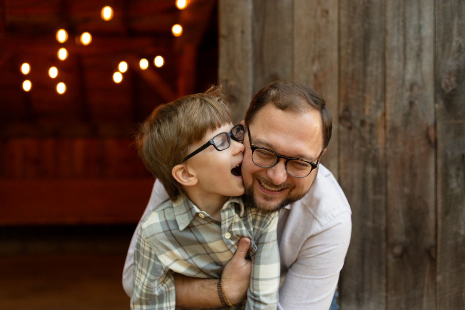 A smiling man in glasses hugs a laughing young boy in glasses and a plaid shirt in front of a rustic wooden barn with string lights.