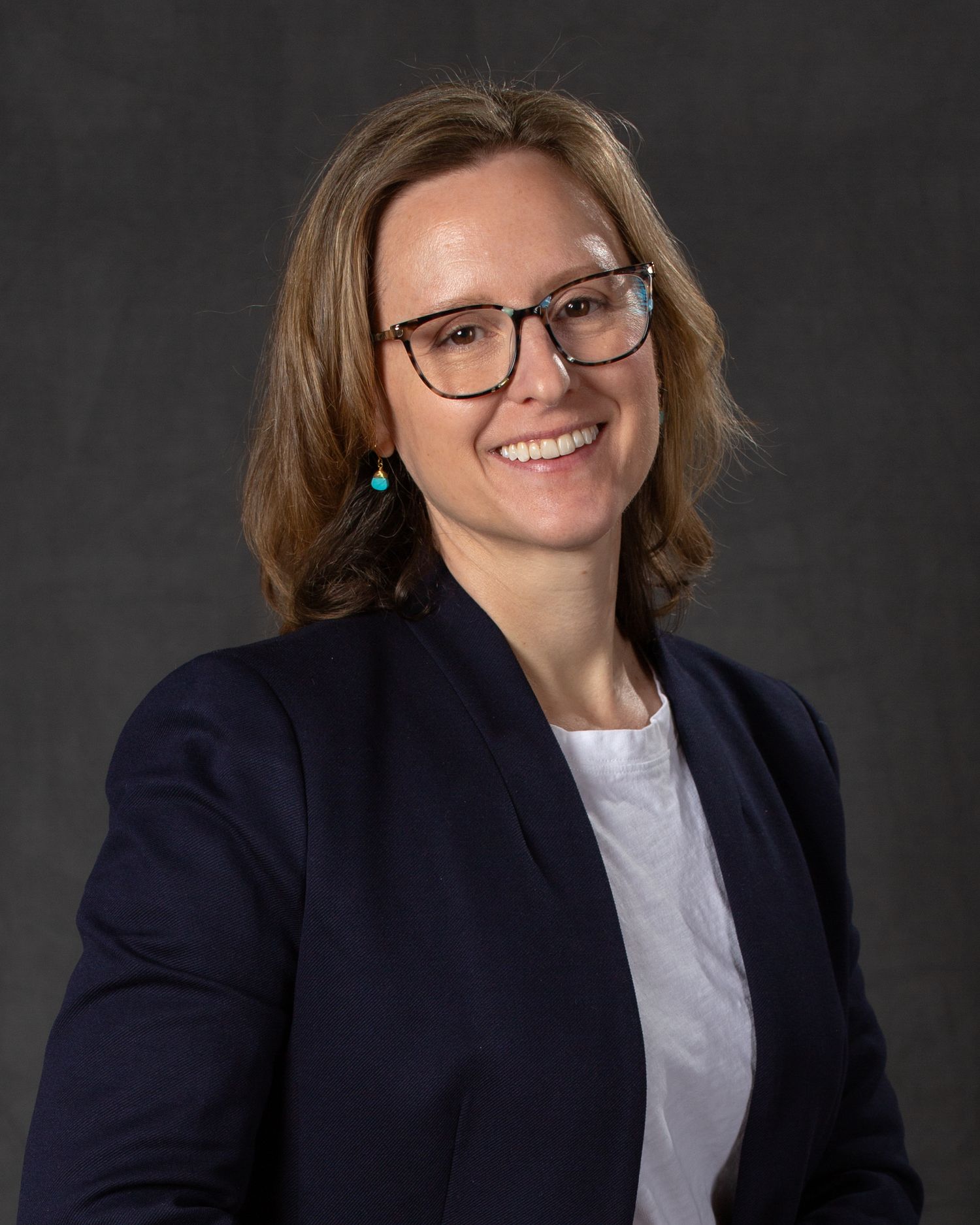 A smiling woman with brown hair and glasses wearing a navy blazer poses for a professional headshot.