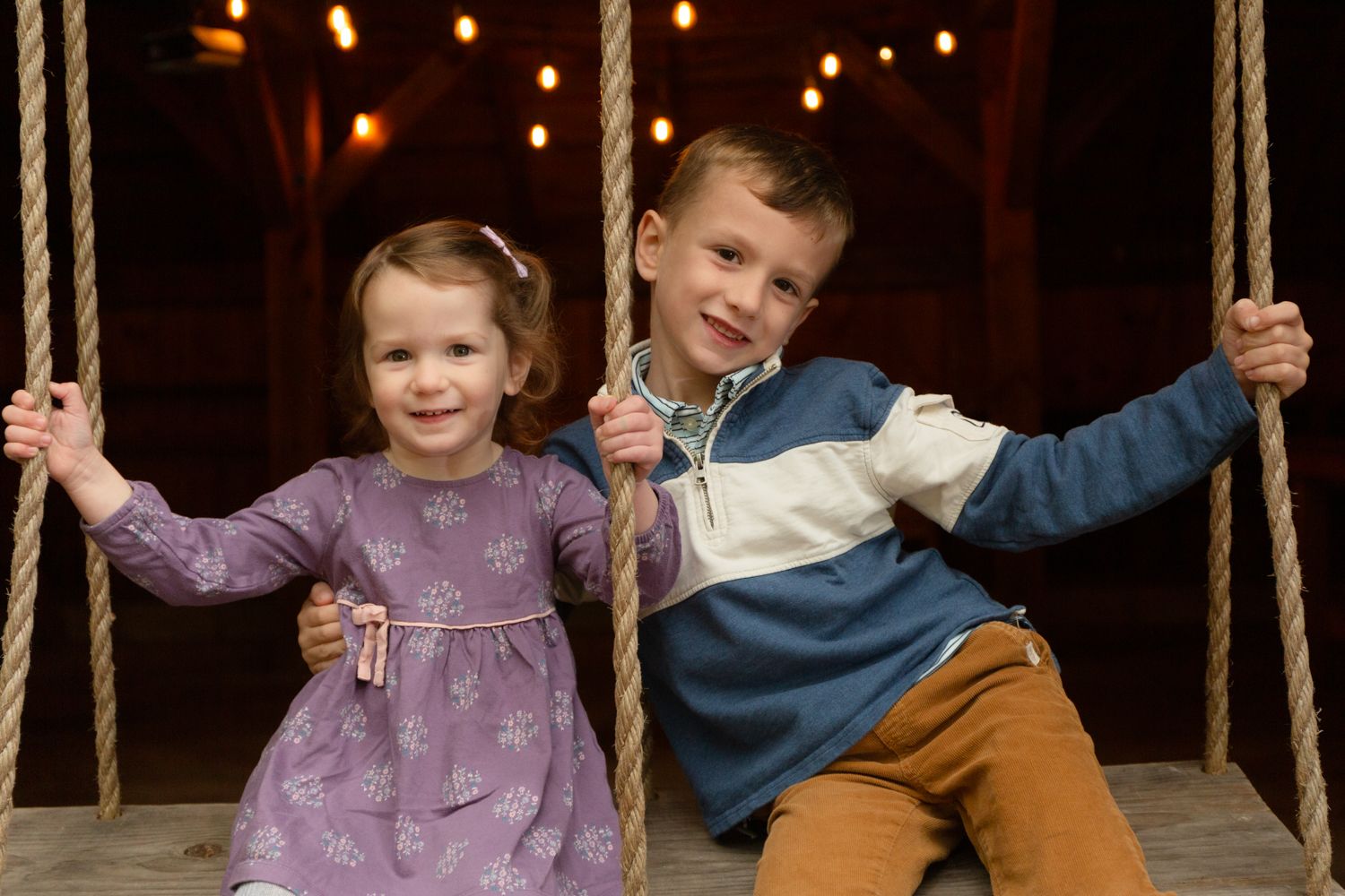 Smiling young girl in a purple dress and a boy in a striped sweater sit together on a rope swing in a warmly lit barn.
