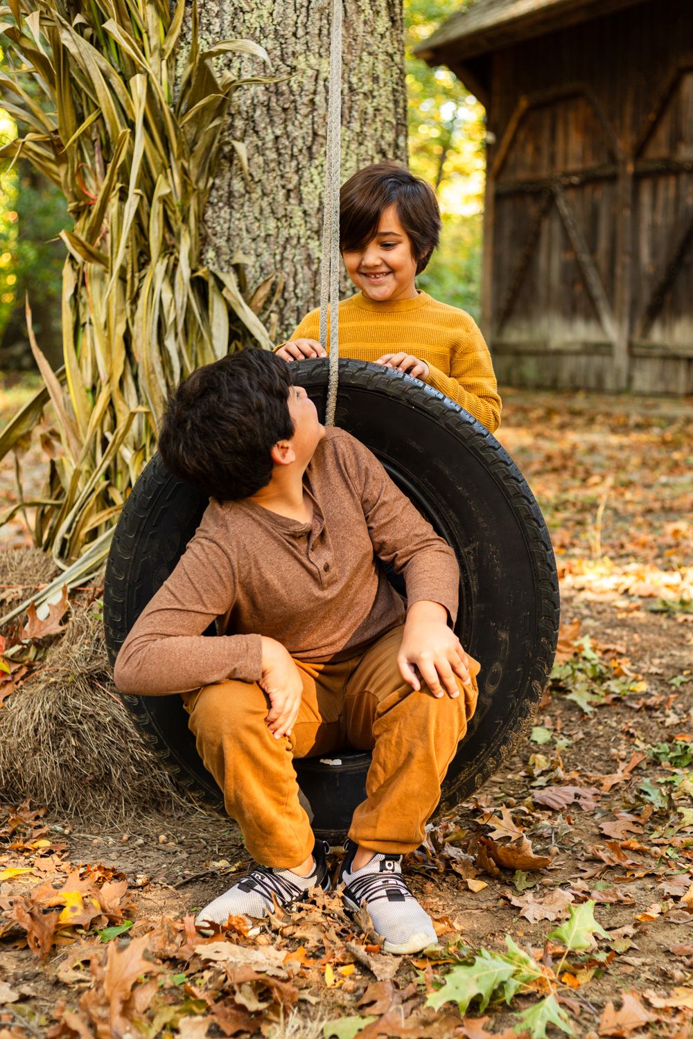 Two boys in autumn outfits play on a tire swing hanging from a tree, surrounded by fallen leaves, corn stalks and an old barn.