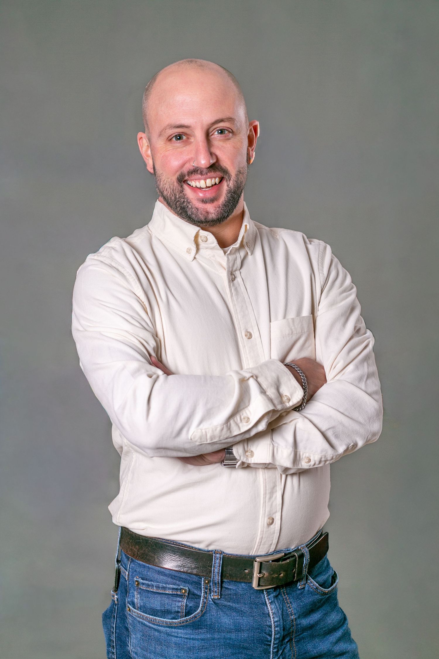 Smiling bald man with beard, arms crossed, wearing white shirt and jeans against gray background.