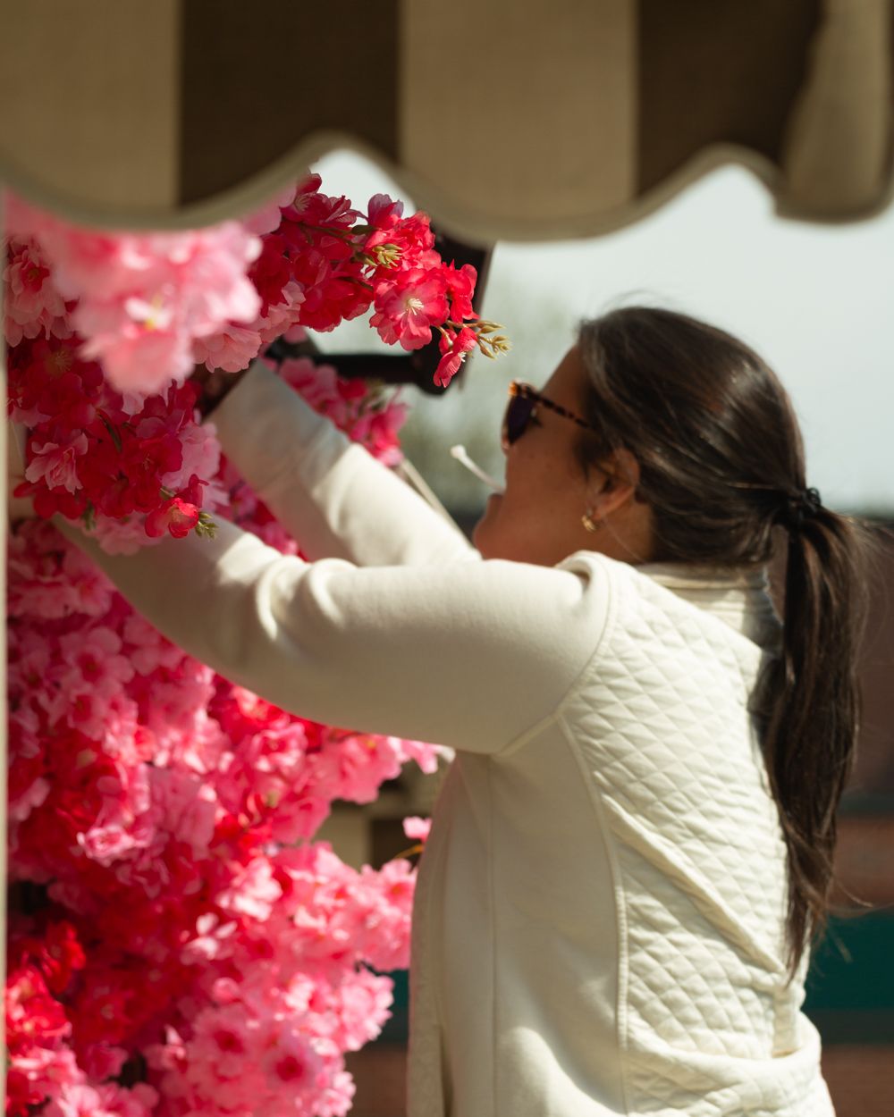 Woman in white quilted jacket arranging pink and red artificial cherry blossom flowers on storefront display.