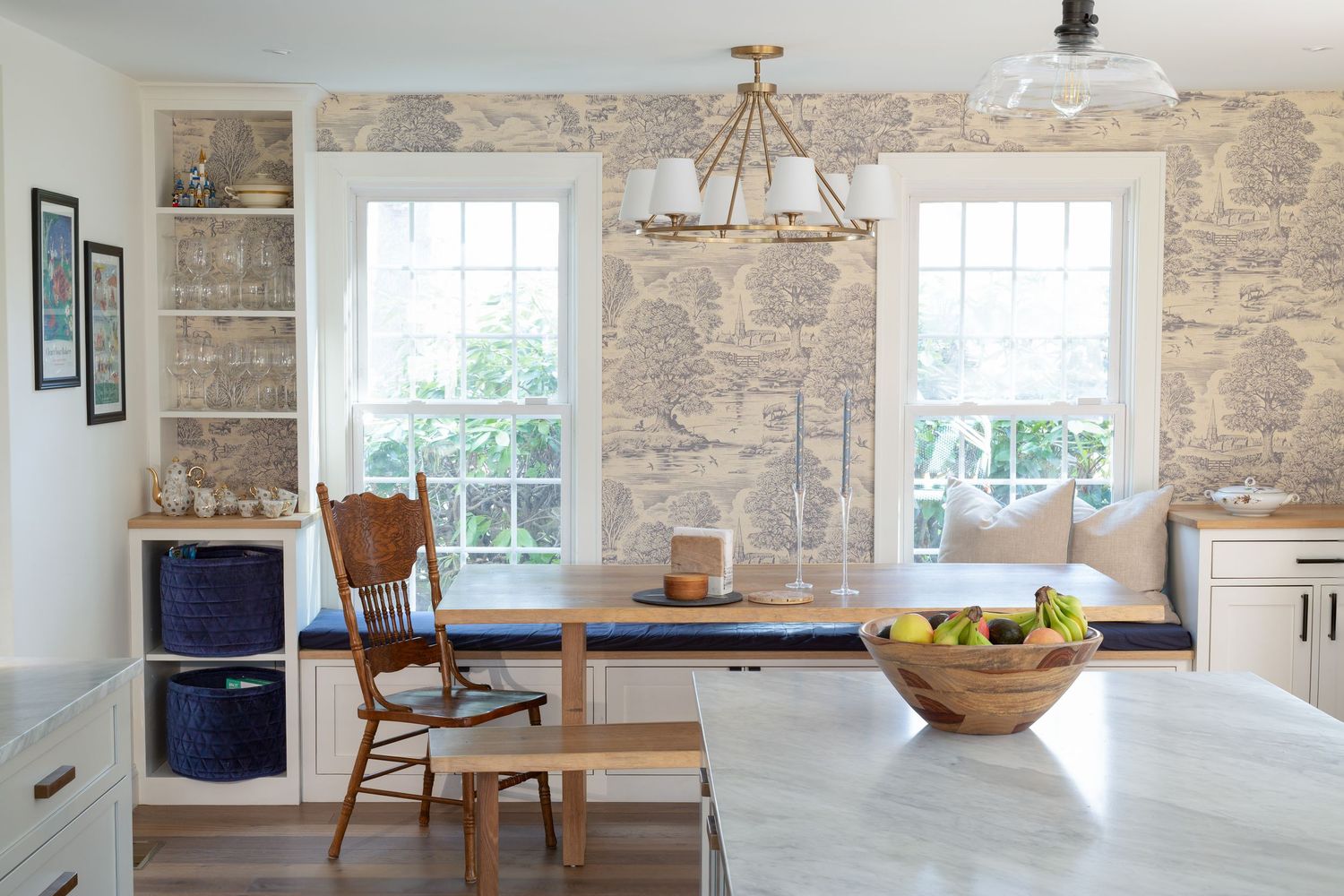Bright breakfast nook with blue built-in bench, wooden table, toile wallpaper, and white shelving in a cozy kitchen.