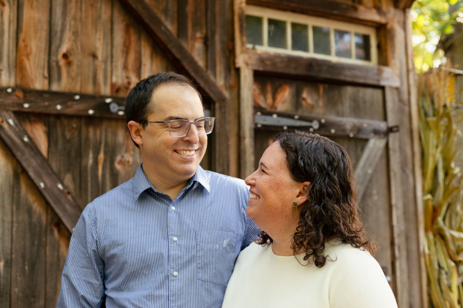 A smiling man with glasses and a woman with dark curly hair gaze warmly at each other in front of a weathered wooden barn door.