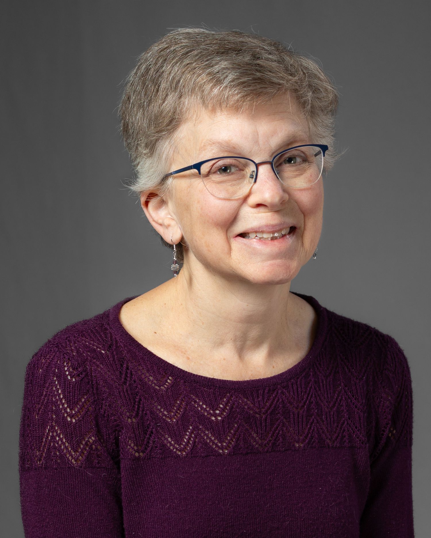 A smiling woman with short gray hair and glasses wearing a purple sweater poses for a headshot.