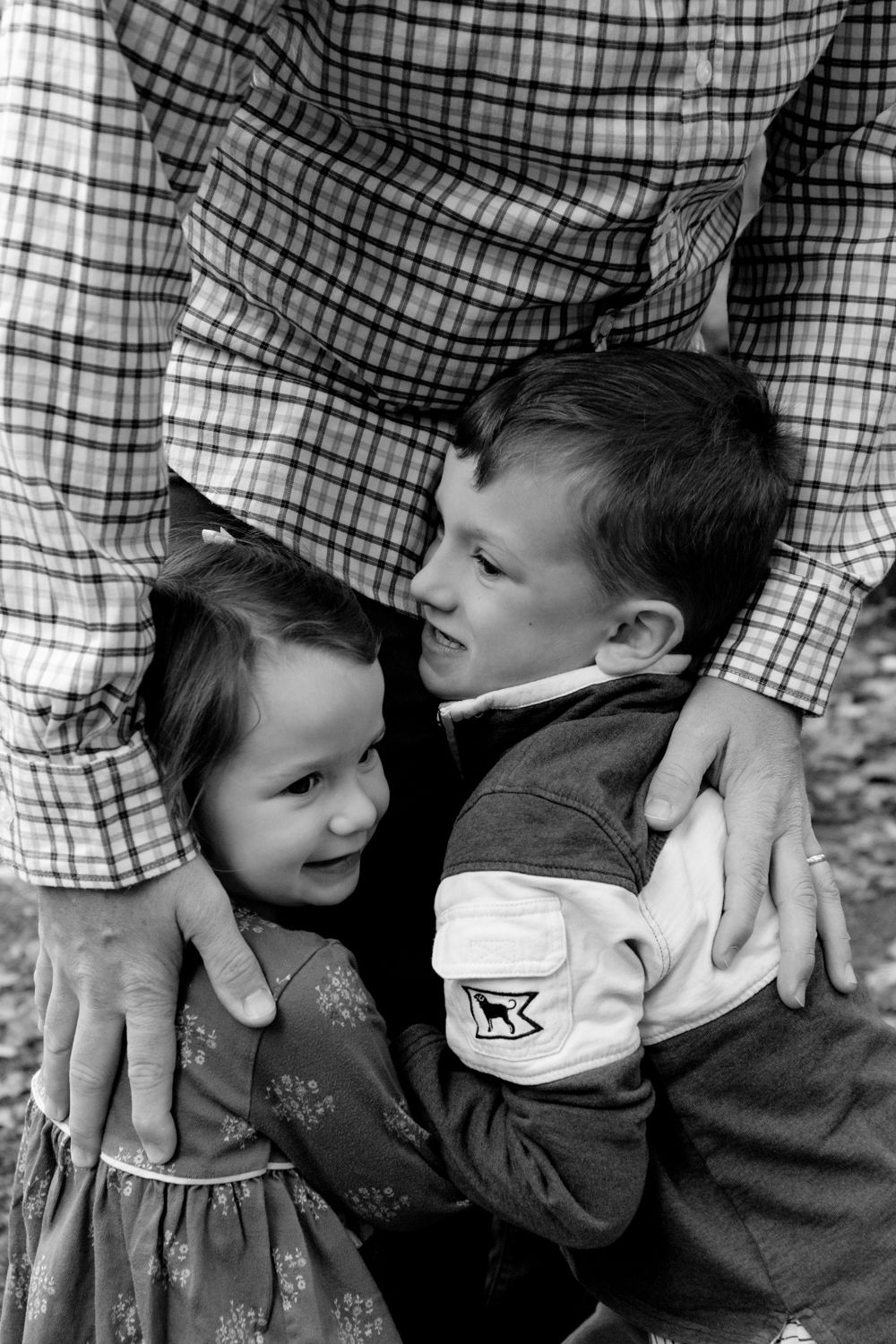 Black and white photo of a young girl and boy hugging close while an adult in a plaid shirt wraps his arms around them.