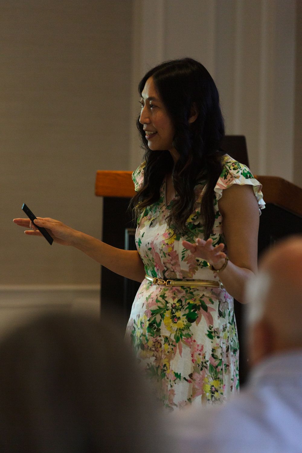 Woman in floral dress presenting to audience while holding a device, smiling confidently during indoor event.
