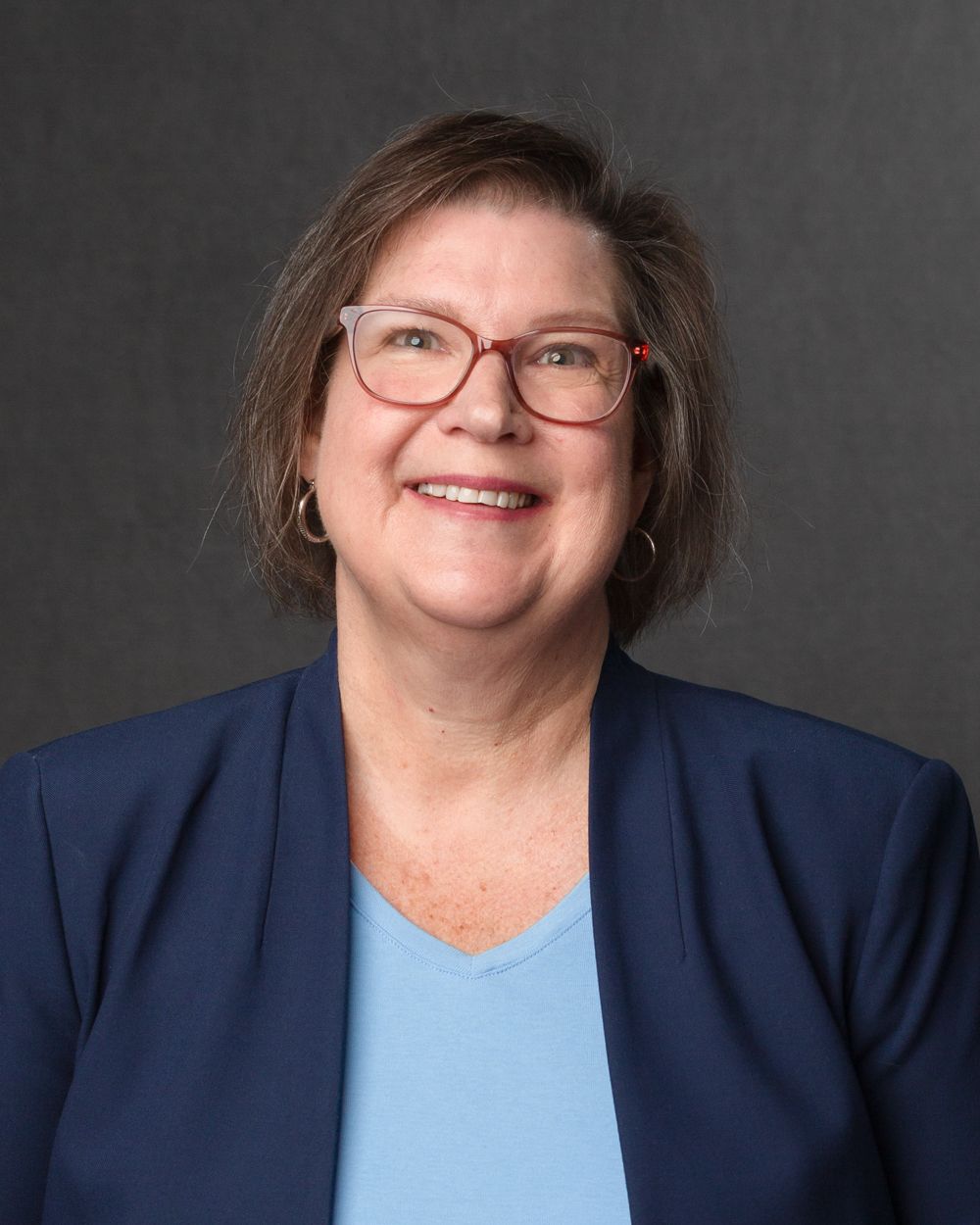Smiling woman with red glasses in navy blazer and blue top on gray background.
