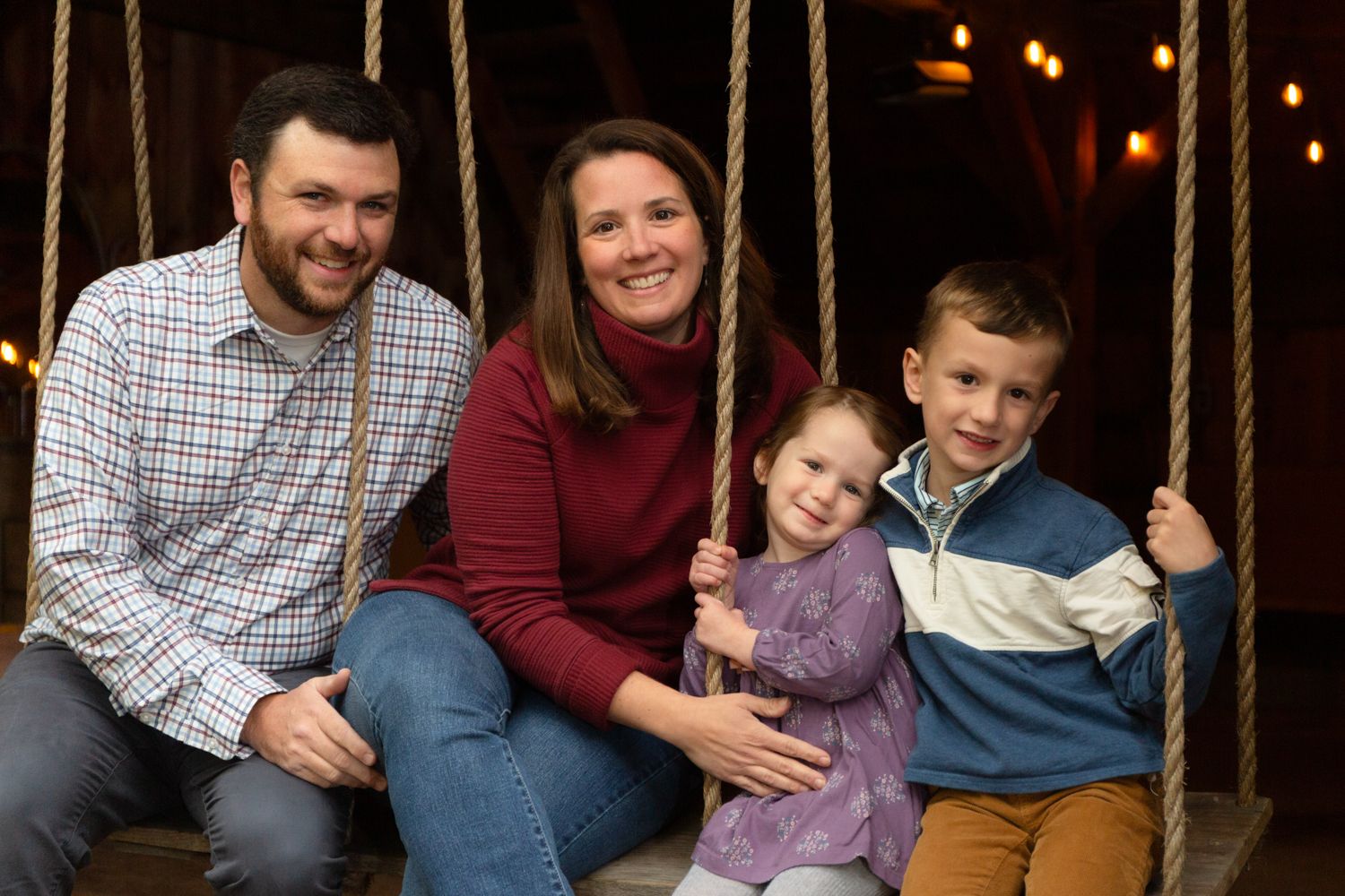 A smiling family of four, two parents and two young children, sits together on a rope swing in a cozy, warmly lit barn.
