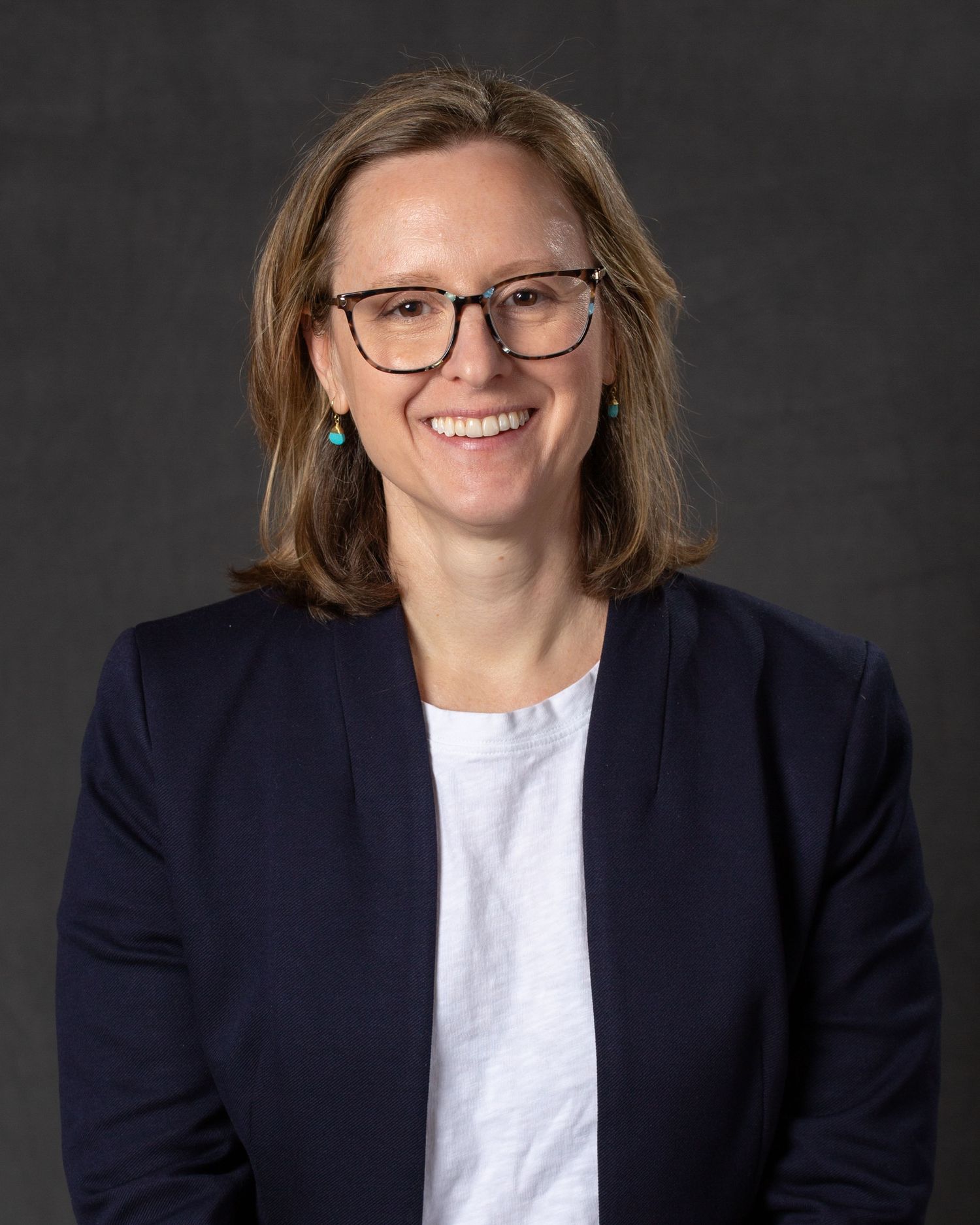 A smiling woman with brown hair and glasses wearing a navy blazer poses for a professional portrait.
