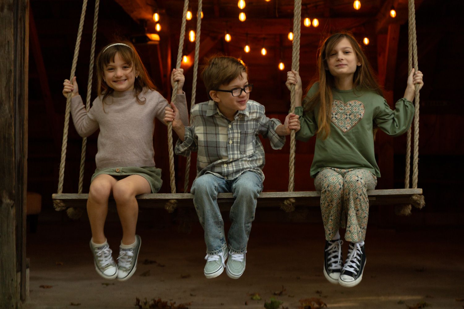 Three smiling children sit side by side on a wooden swing at dusk, with warm string lights glowing in the background.