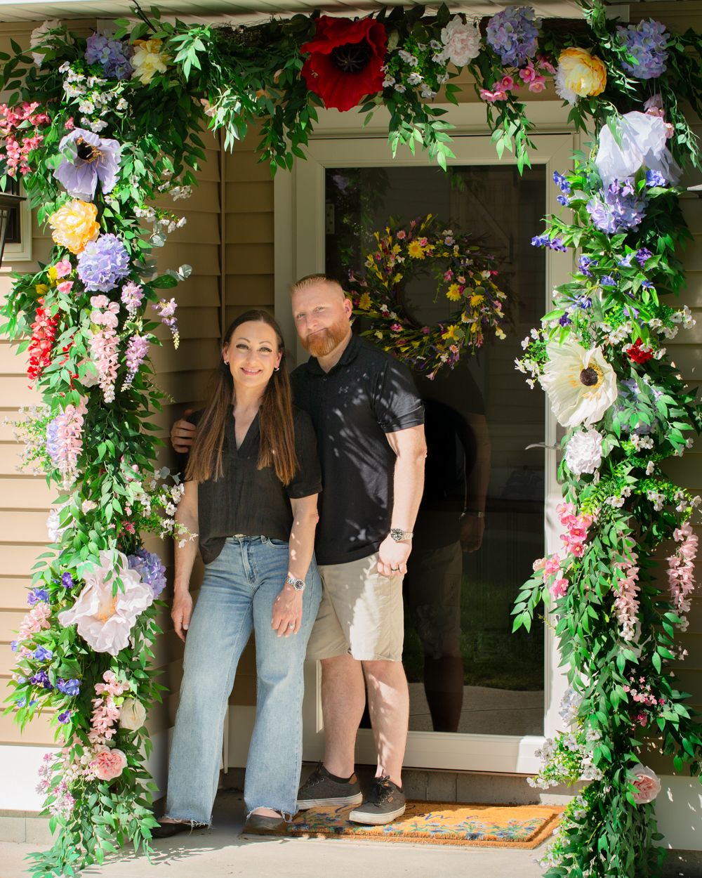 Smiling couple posing under a lush colorful floral arch at their home's front entrance.