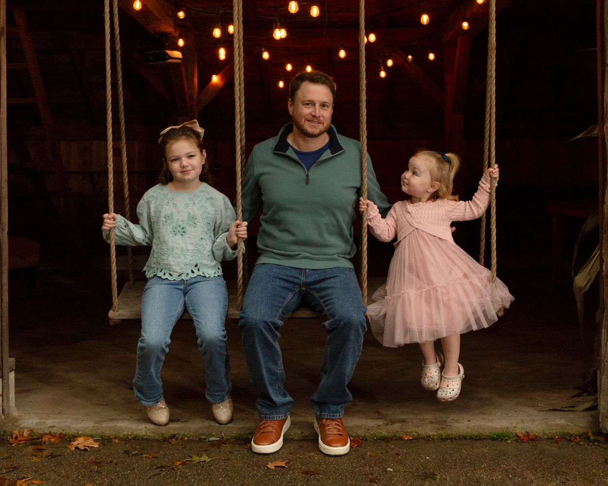 A man in a green sweater sits on a wooden swing between two young girls in a cozy barn lit by warm string lights.