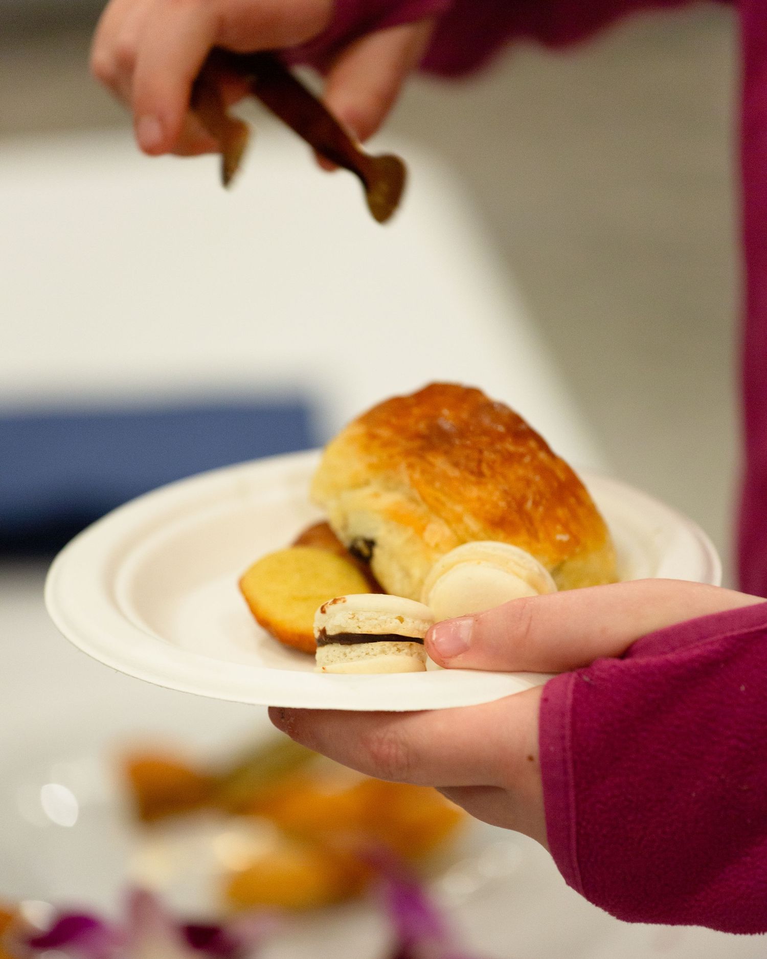 Hands holding white plate with golden baked scone and sliced fruit at food event.