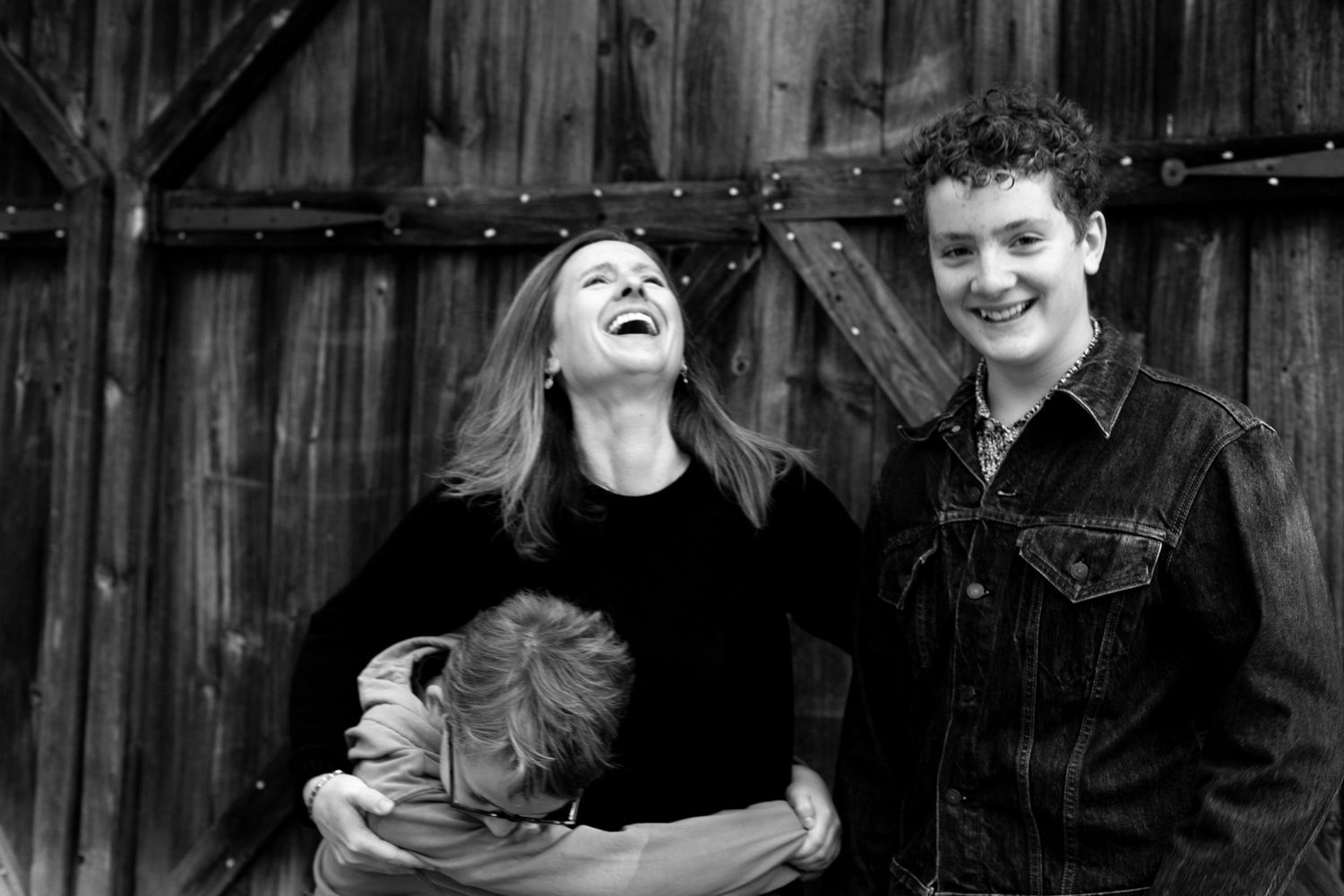 Black and white photo of a laughing woman hugging a child while a smiling teen in a denim jacket stands by a weathered wooden barn door.