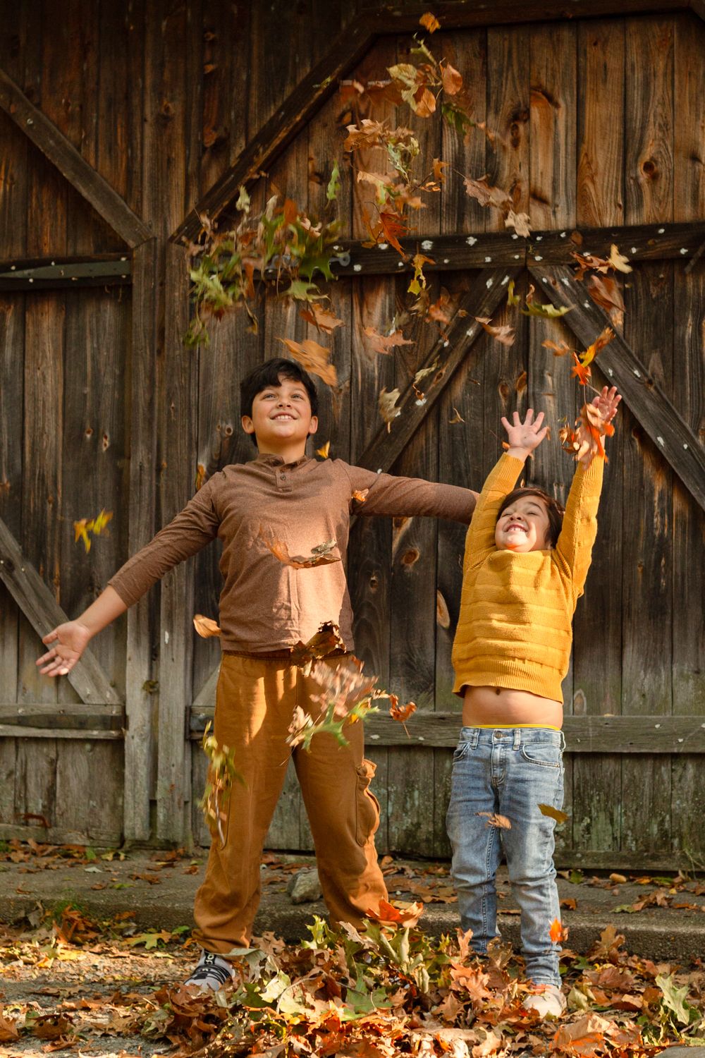 Two smiling children toss colorful autumn leaves into the air in front of a rustic wooden barn door on a sunny fall day.
