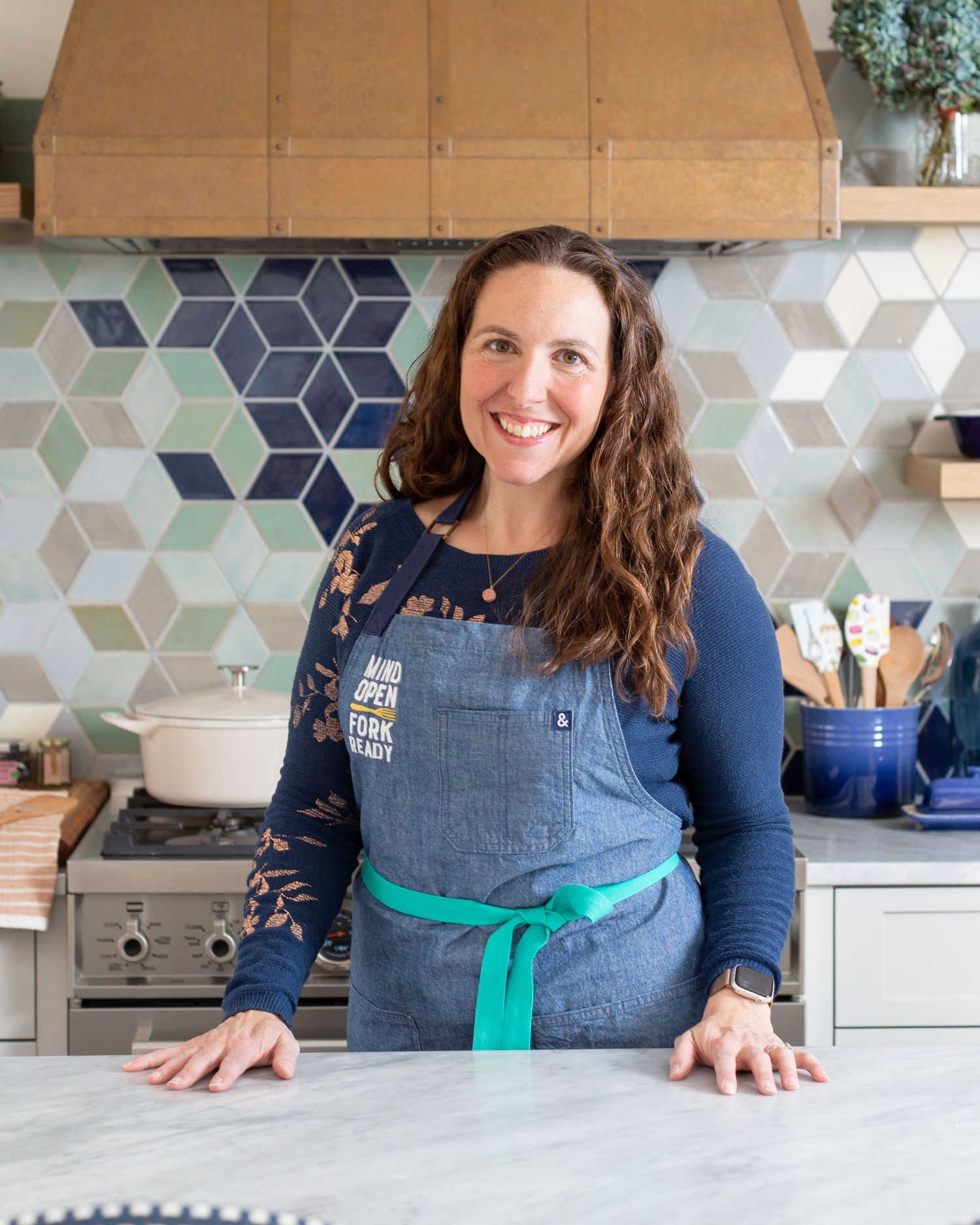 Woman in denim apron leans on marble kitchen counter with multicolor diamond tile backsplash behind her.