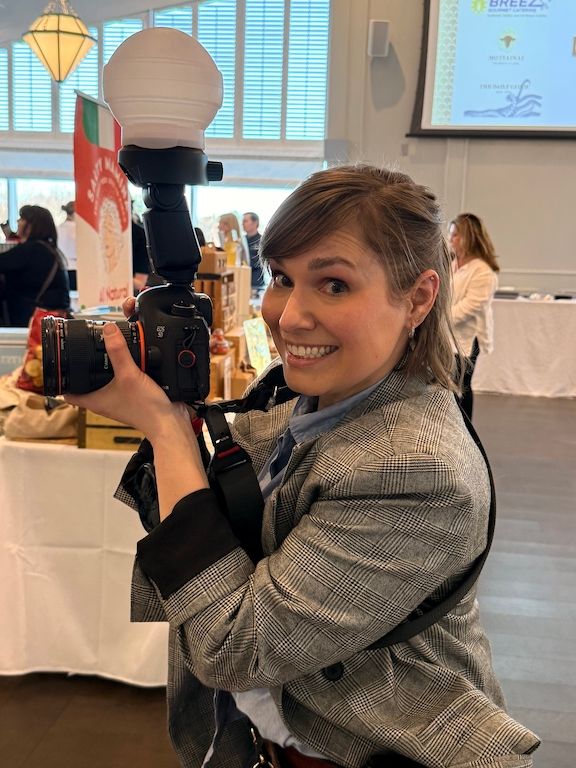 Smiling female photographer holding a DSLR camera with flash at an indoor event.