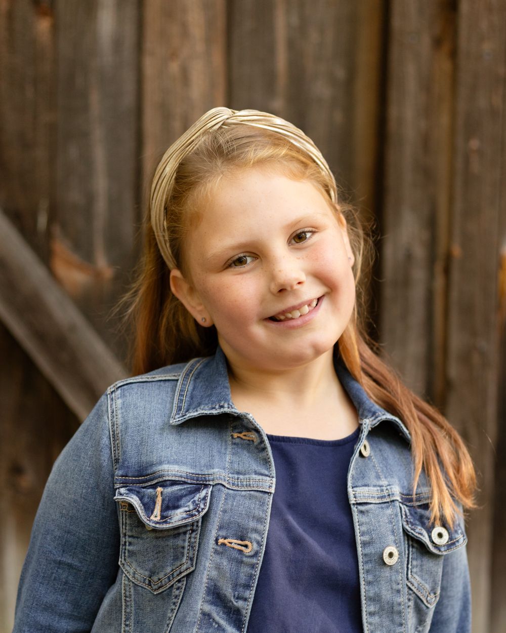A smiling young girl with long red hair, a gold headband and a denim jacket stands before a rustic wooden barn wall.