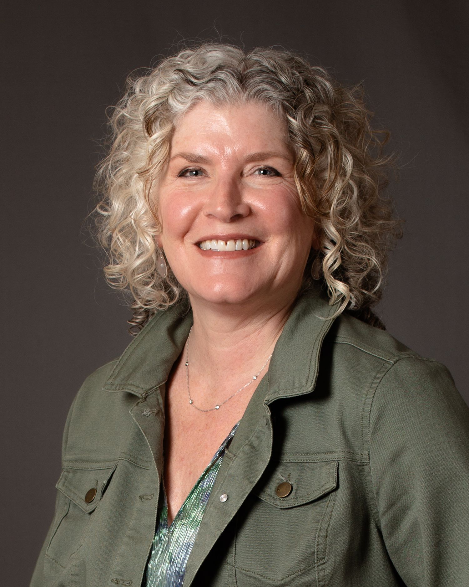 A smiling woman with curly gray hair wearing a green jacket poses for a professional headshot.