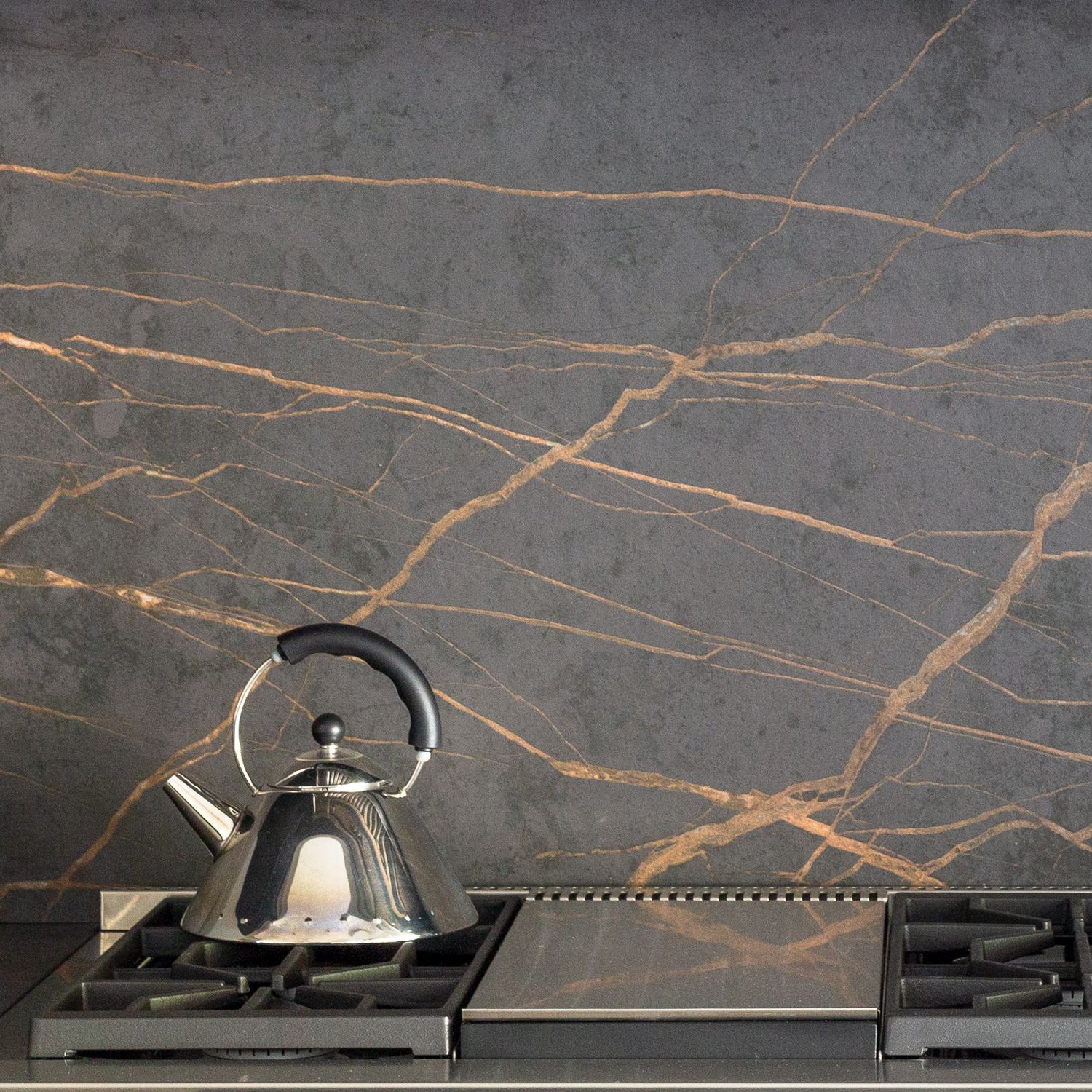 Dark gray marble backsplash with gold veining behind a stovetop with a stainless steel kettle.