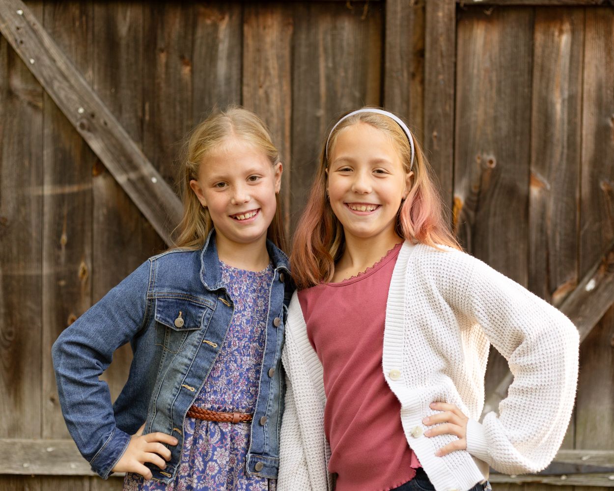 Two smiling girls in a denim jacket and a white cardigan pose side by side in front of a rustic weathered wooden barn door.