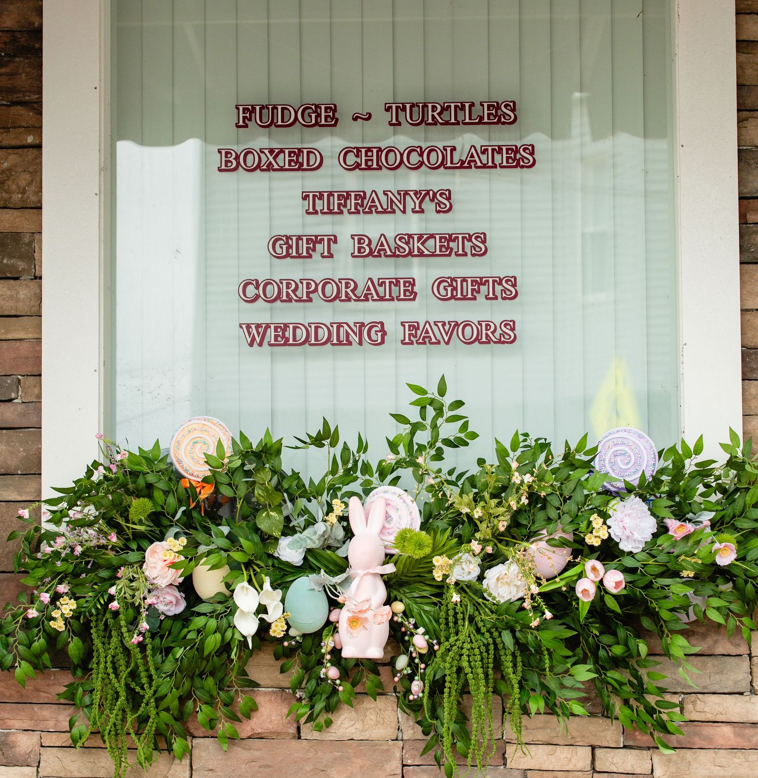 Window display of a candy shop advertising fudge, turtles, boxed chocolates, and gift baskets with spring floral decorations.