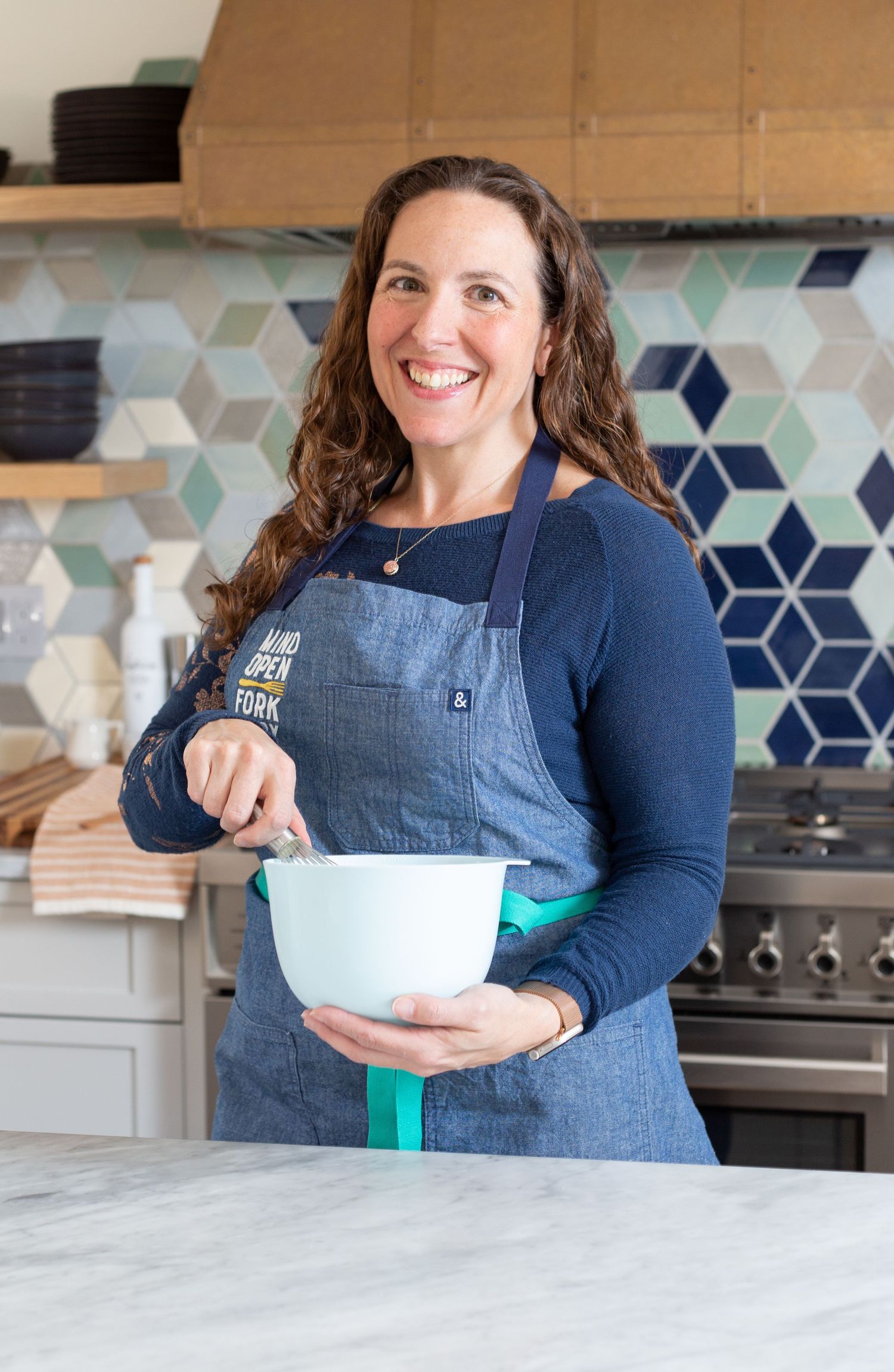 Smiling woman in denim apron mixing ingredients in a white bowl in a modern kitchen.