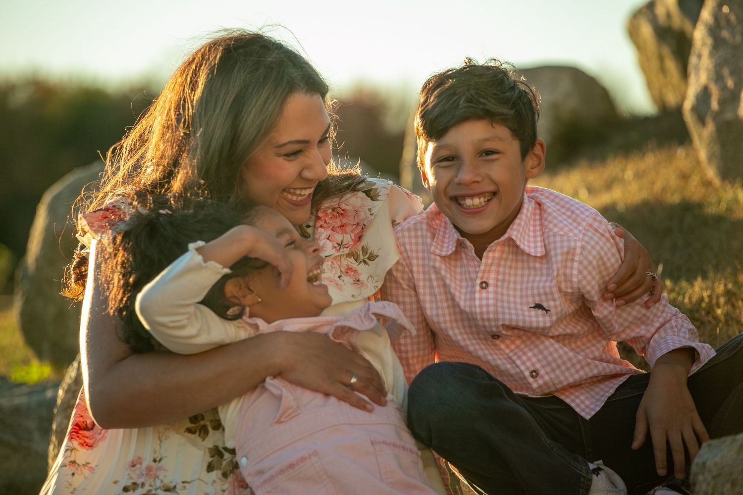 A mother hugs her two young children outdoors at golden hour, all smiling warmly in a sunny field setting.
