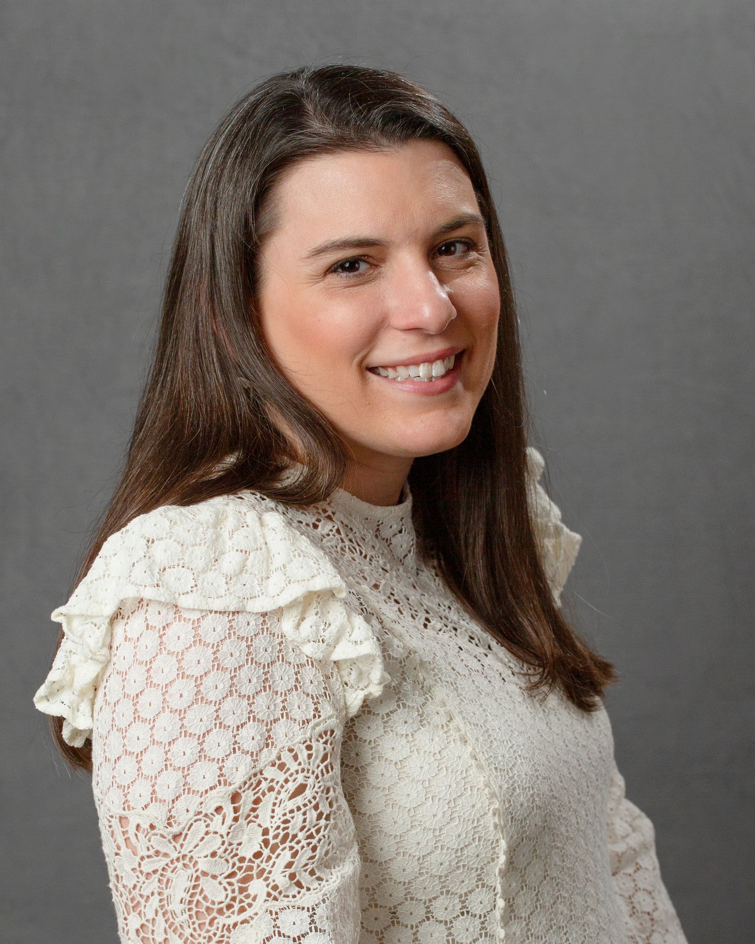 Smiling brunette woman in white lace top with ruffled shoulders against gray background.