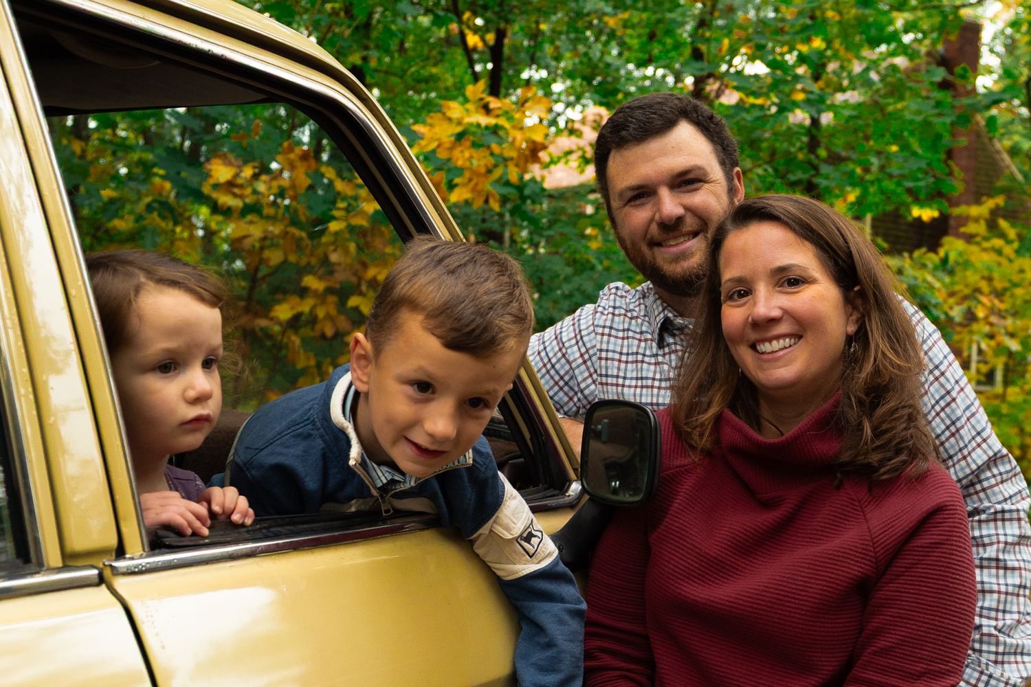 Smiling family of four with two young children and two parents poses by a yellow vintage car surrounded by colorful autumn foliage.