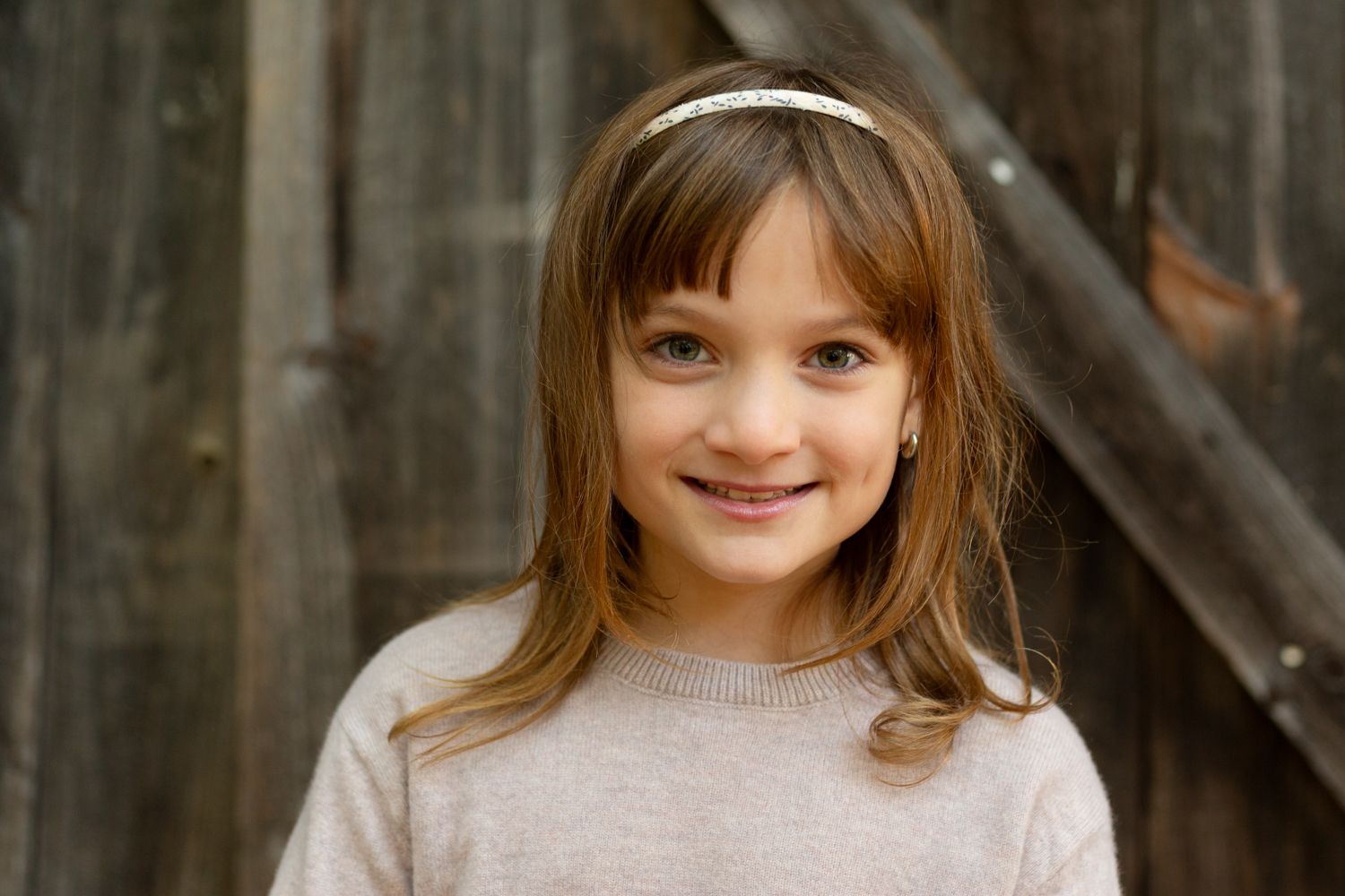 Smiling young girl with long brown hair and a white headband wearing a cream knit sweater in front of a rustic weathered wooden barn door.