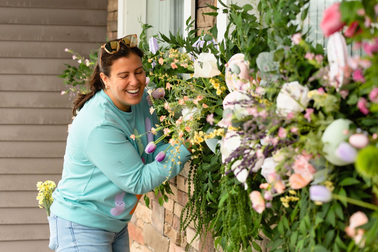 Cheerful woman in teal sweatshirt laughing while arranging a pastel spring floral arrangement.