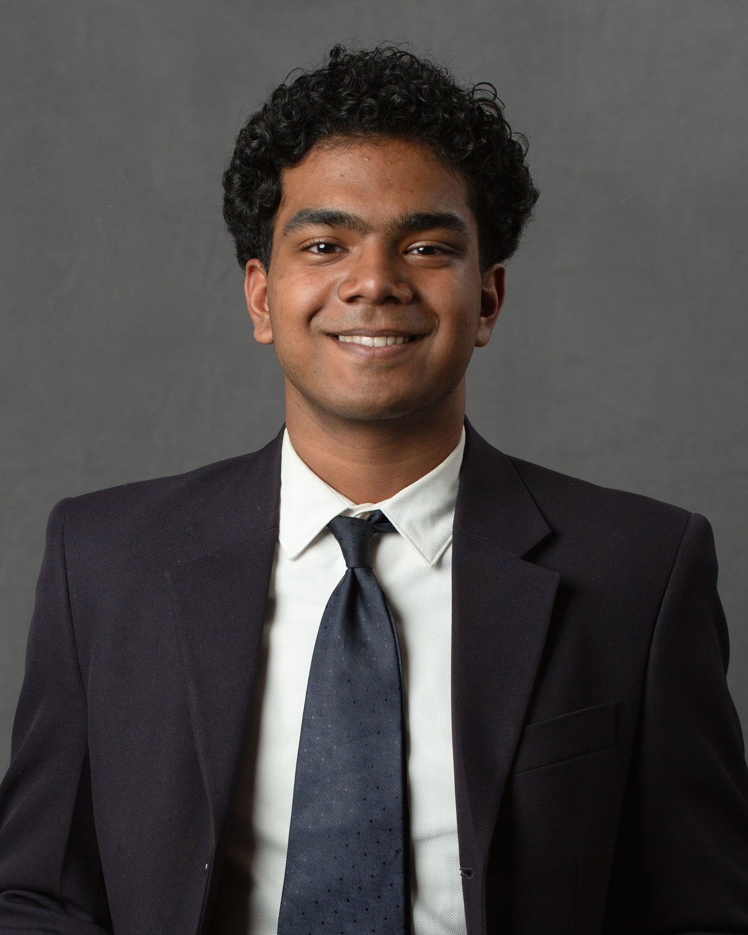 Young man in dark suit and tie smiling for professional headshot on gray background.