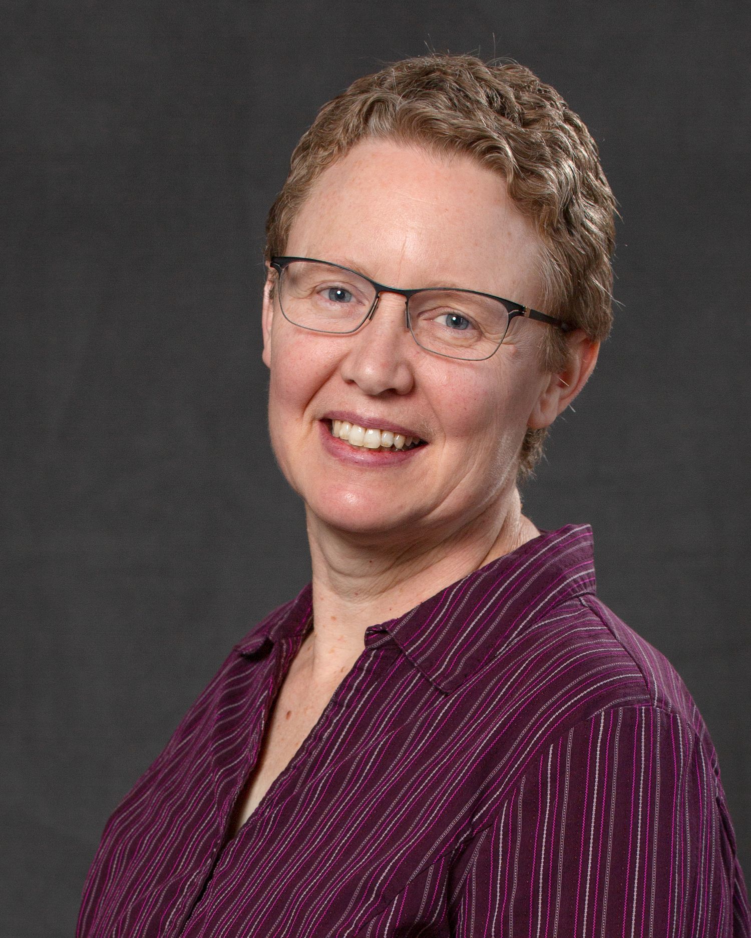 A smiling woman with short blonde hair and glasses wearing a purple striped shirt in a headshot.
