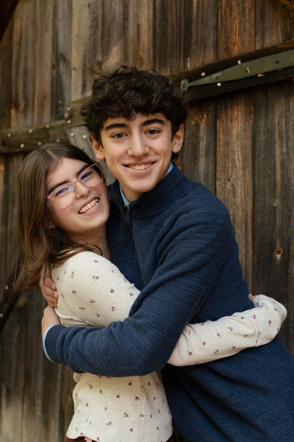 A smiling teenage boy in a navy zip sweater hugs a girl with glasses in front of a rustic wooden barn door.