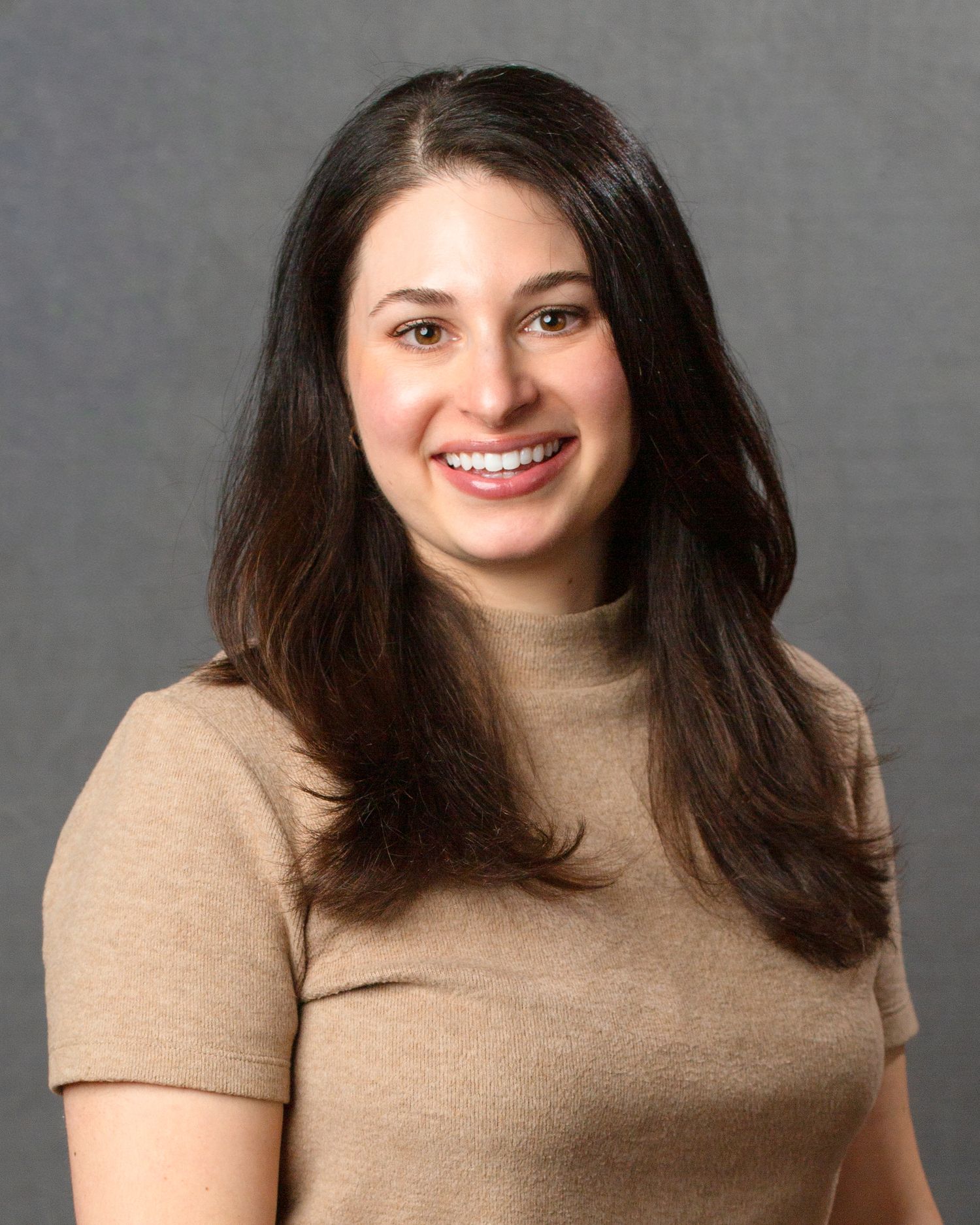 Young brunette woman in tan turtleneck sweater posing for professional headshot on gray background.