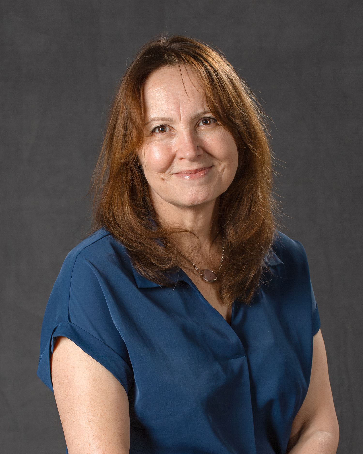A smiling woman with auburn hair wearing a teal blue top poses for a professional headshot.