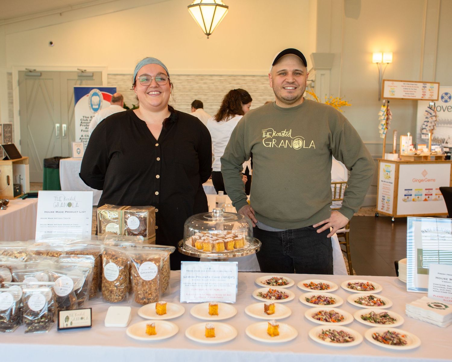 Two vendors smiling behind a granola display table at an indoor food market.