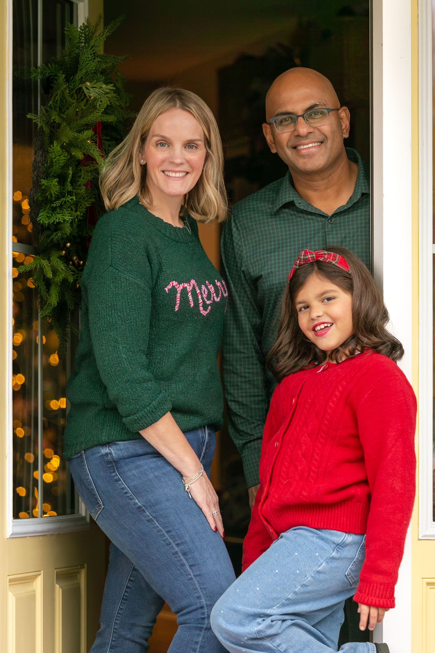 Family of three posing at front door with Christmas wreath, wearing festive green and red holiday outfits.