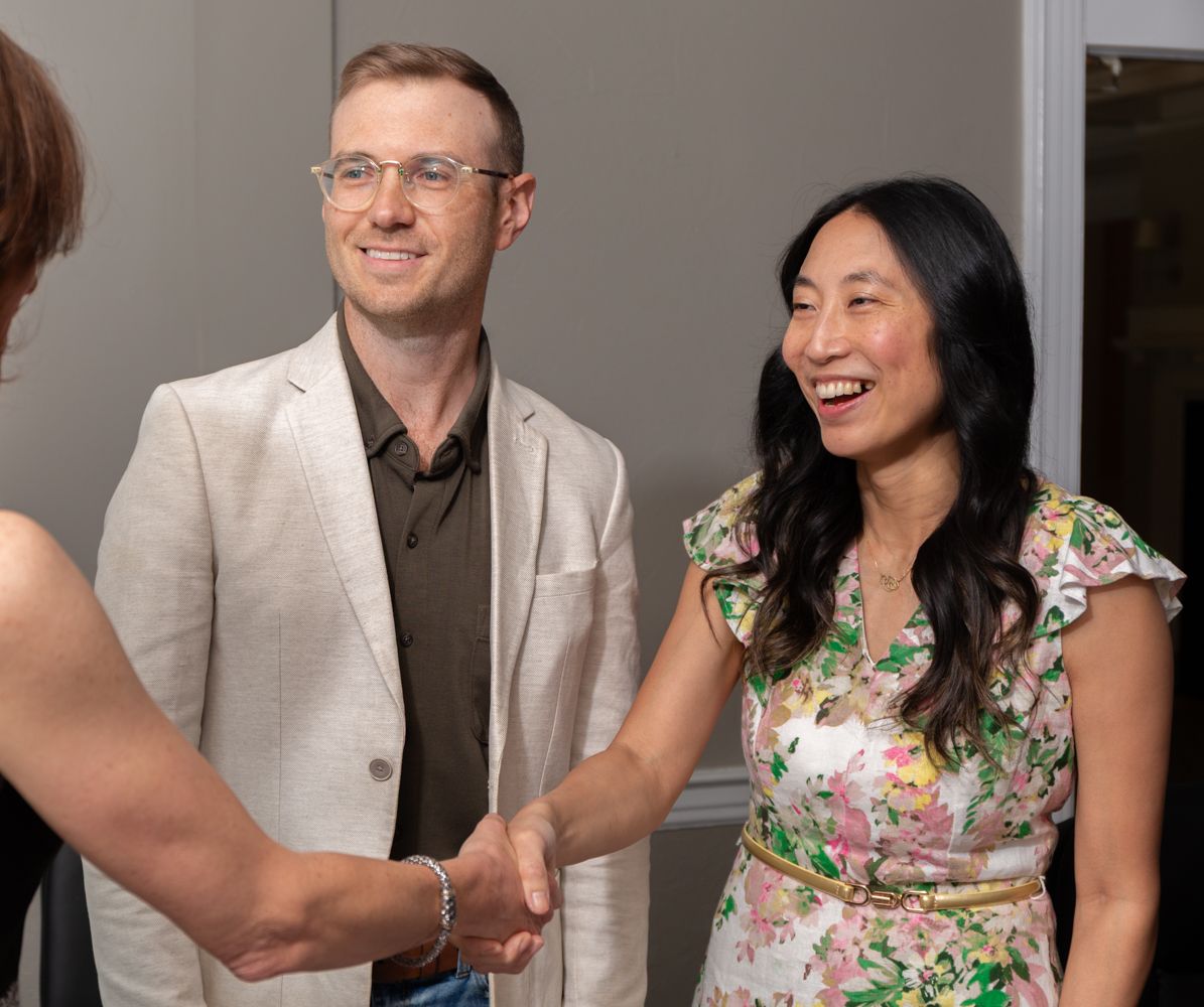 Man in beige blazer and woman in floral dress laugh while shaking hands at a networking event.