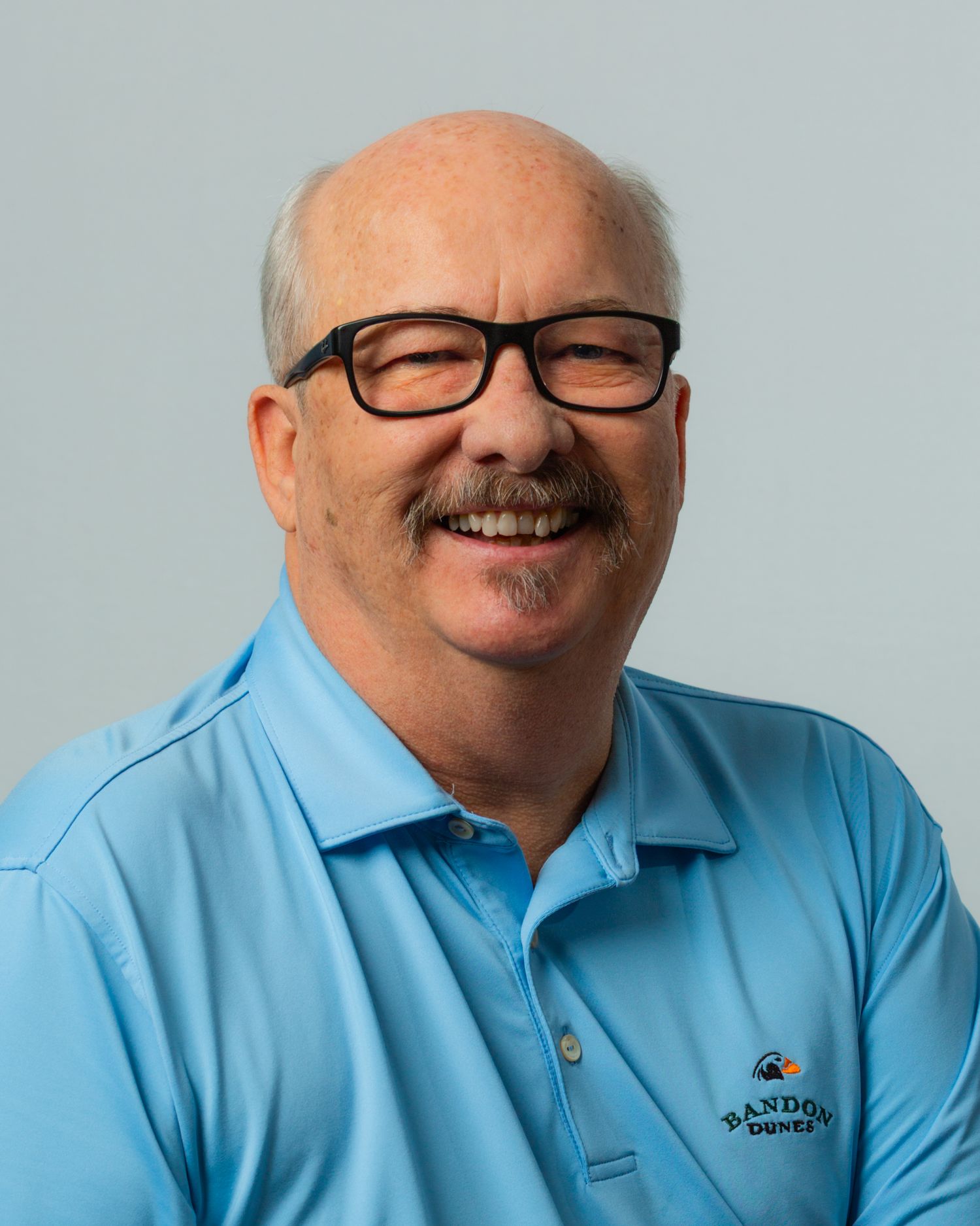 Friendly older man with mustache and glasses wearing a light blue polo shirt, smiling.