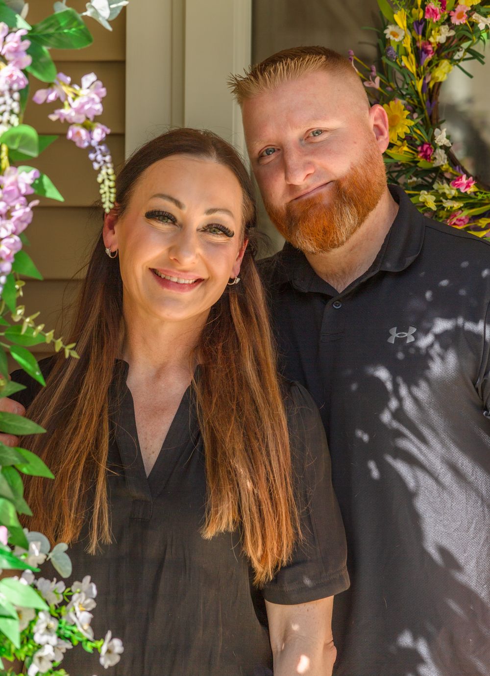 Smiling couple posing together among purple wisteria and yellow flowers outside a home.
