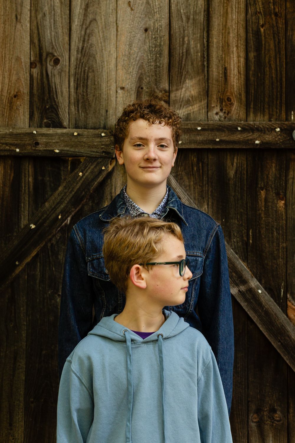 A tall teenager in a denim jacket stands behind a younger boy with glasses in a blue hoodie against a weathered barn door.