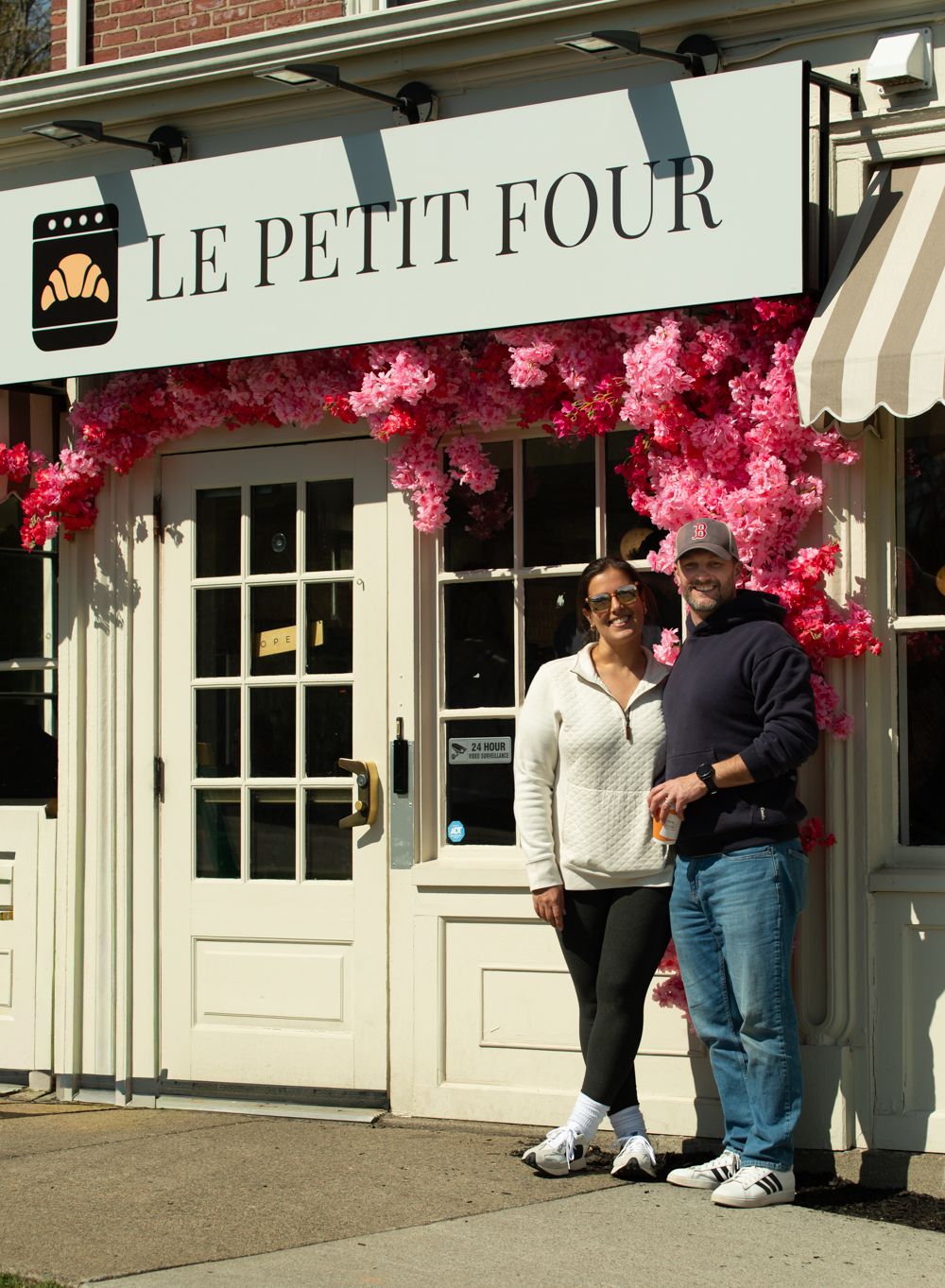 Couple smiling outside Le Petit Four bakery with pink floral decorations.