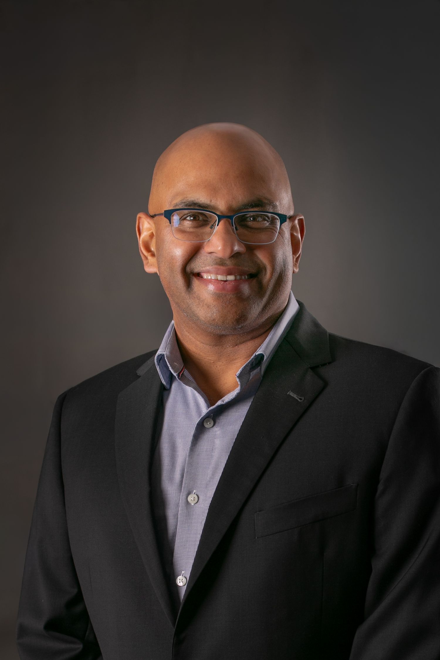 Professional bald man wearing glasses and black blazer smiling against dark background.