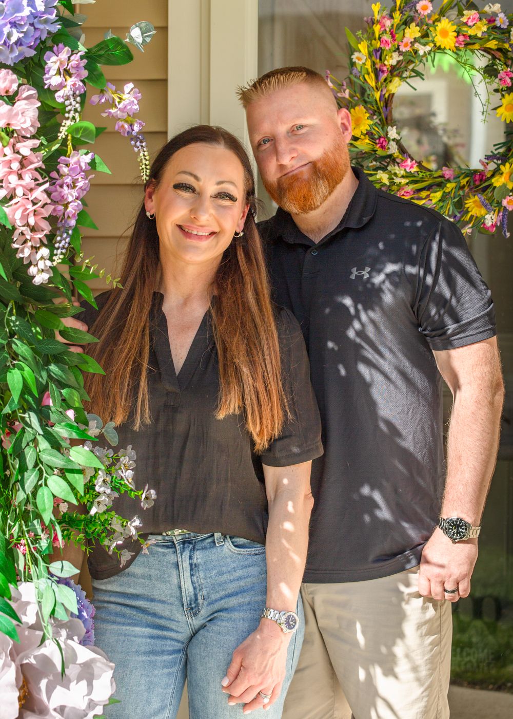 Smiling couple posing together outdoors in front of a colorful spring floral arch decoration.